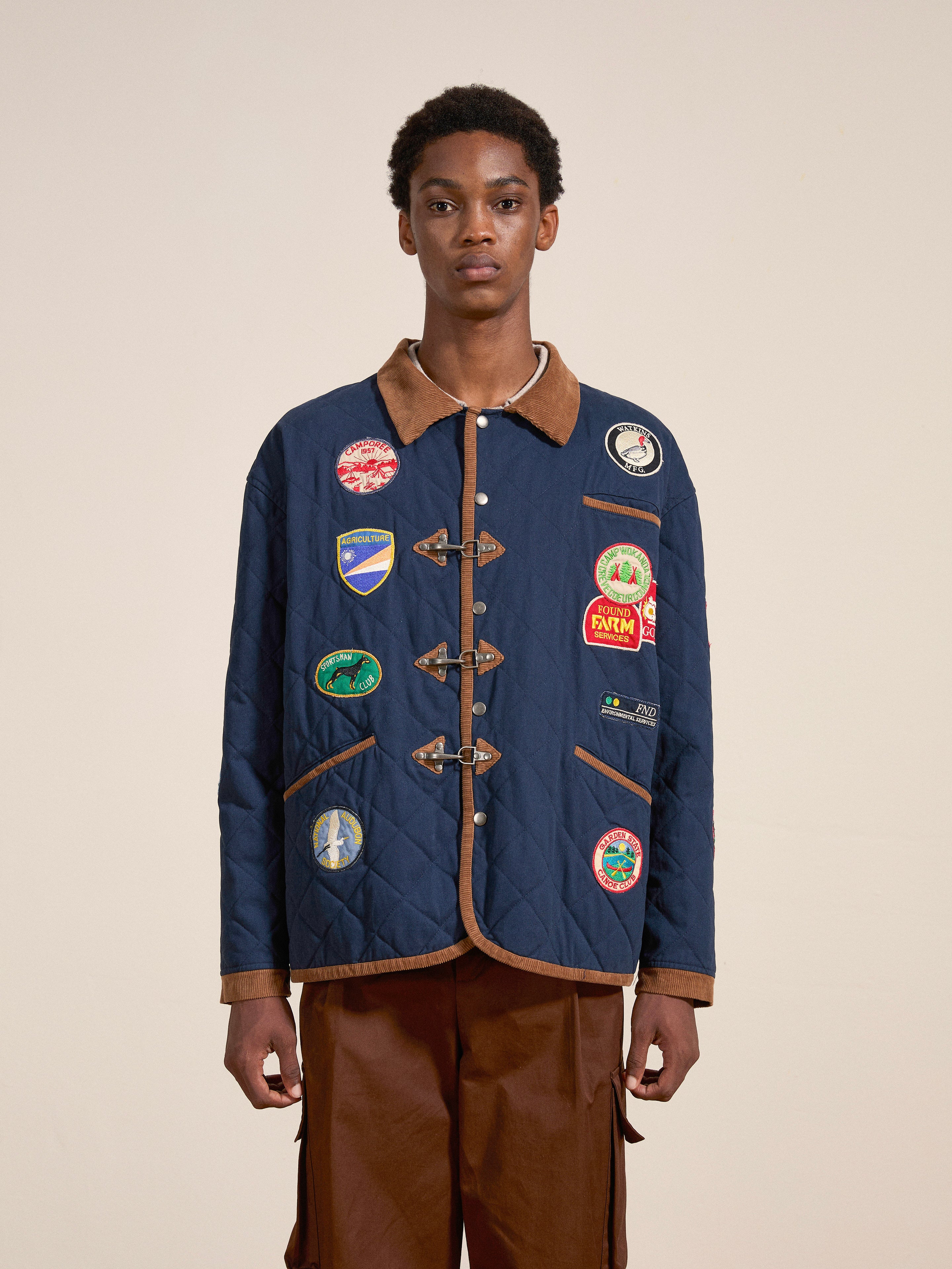 A person stands wearing the FOUND Farmstead Quilt Patch Jacket—an eye-catching blue quilted jacket embellished with vibrant patches reminiscent of vintage hunting competition jackets and brown trim—paired with brown pants, against a neutral background.