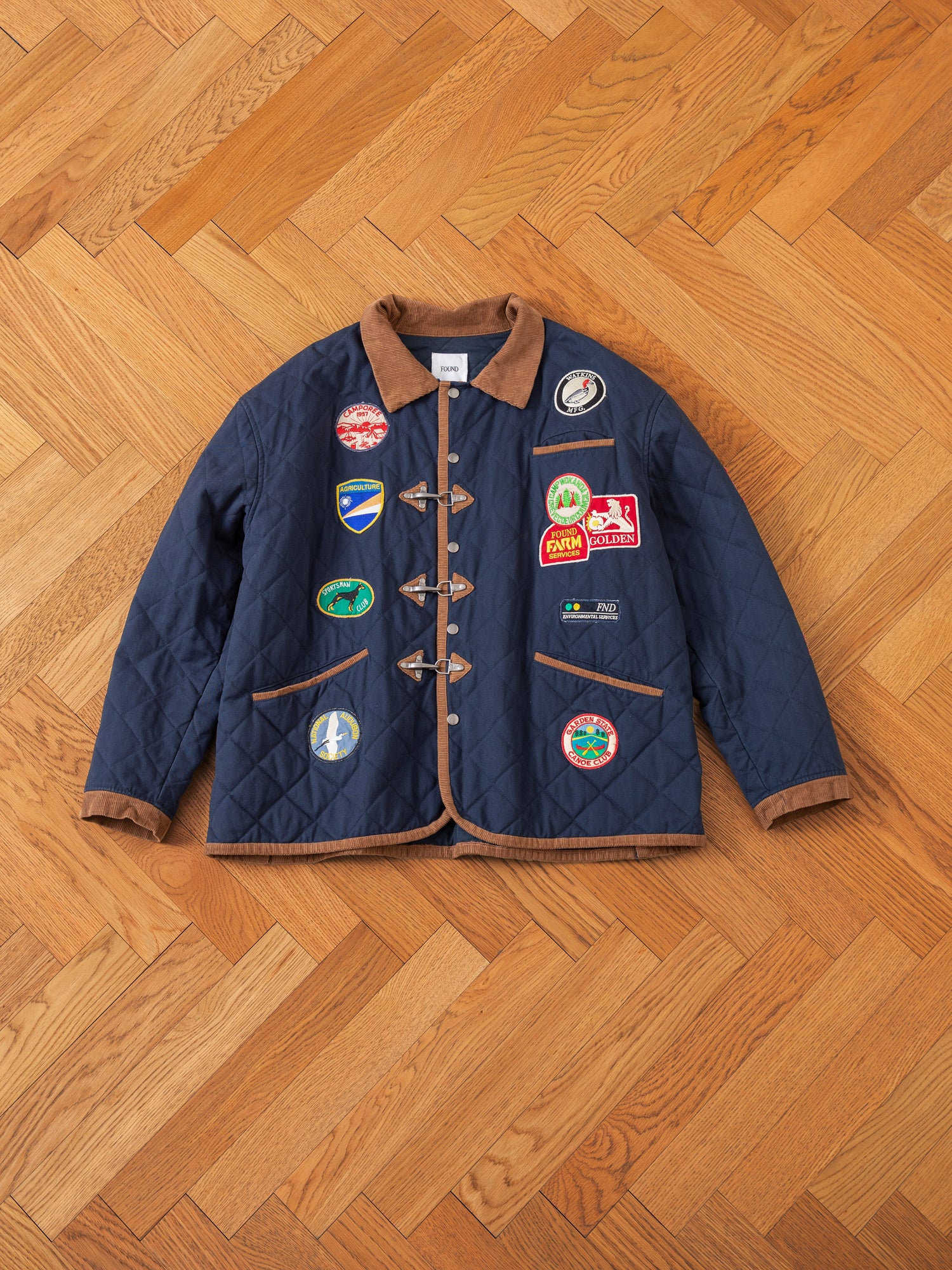 The FOUND Farmstead Quilt Patch Jacket in blue, accented with a brown collar and edging, showcases an array of colorful patches on the front. Reminiscent of vintage hunting competition jackets, it is set against a wood-patterned floor, exuding the rustic charm of farm stands.