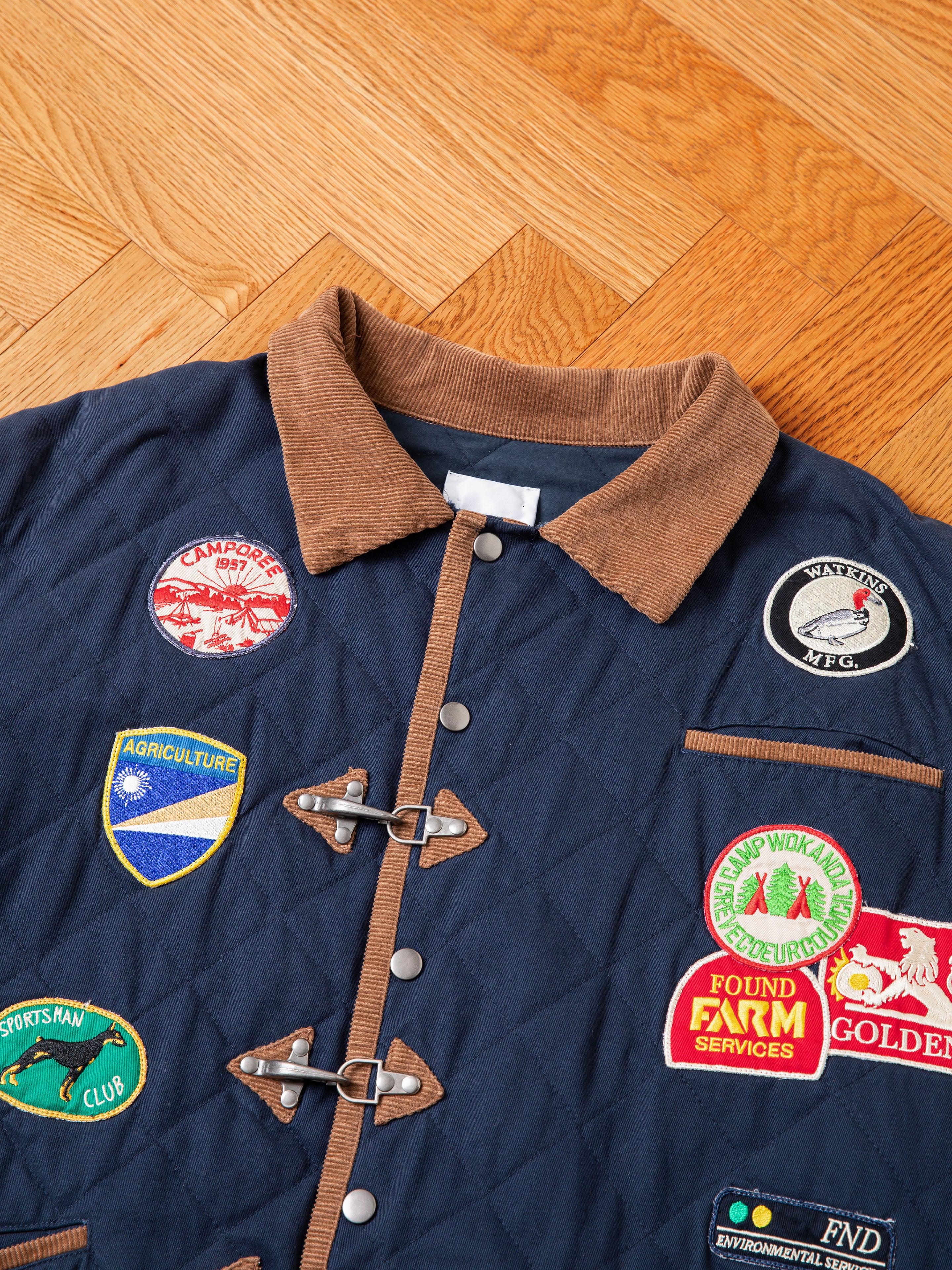 The Farmstead Quilt Patch Jacket by FOUND, featuring a blue quilted design with a brown corduroy collar and wooden toggle buttons, is laid out on a wooden floor. Adorned with various colorful patches related to agriculture, camps, and sports, this jacket evokes the charm of vintage hunting competition jackets—ideal for farm stand enthusiasts.