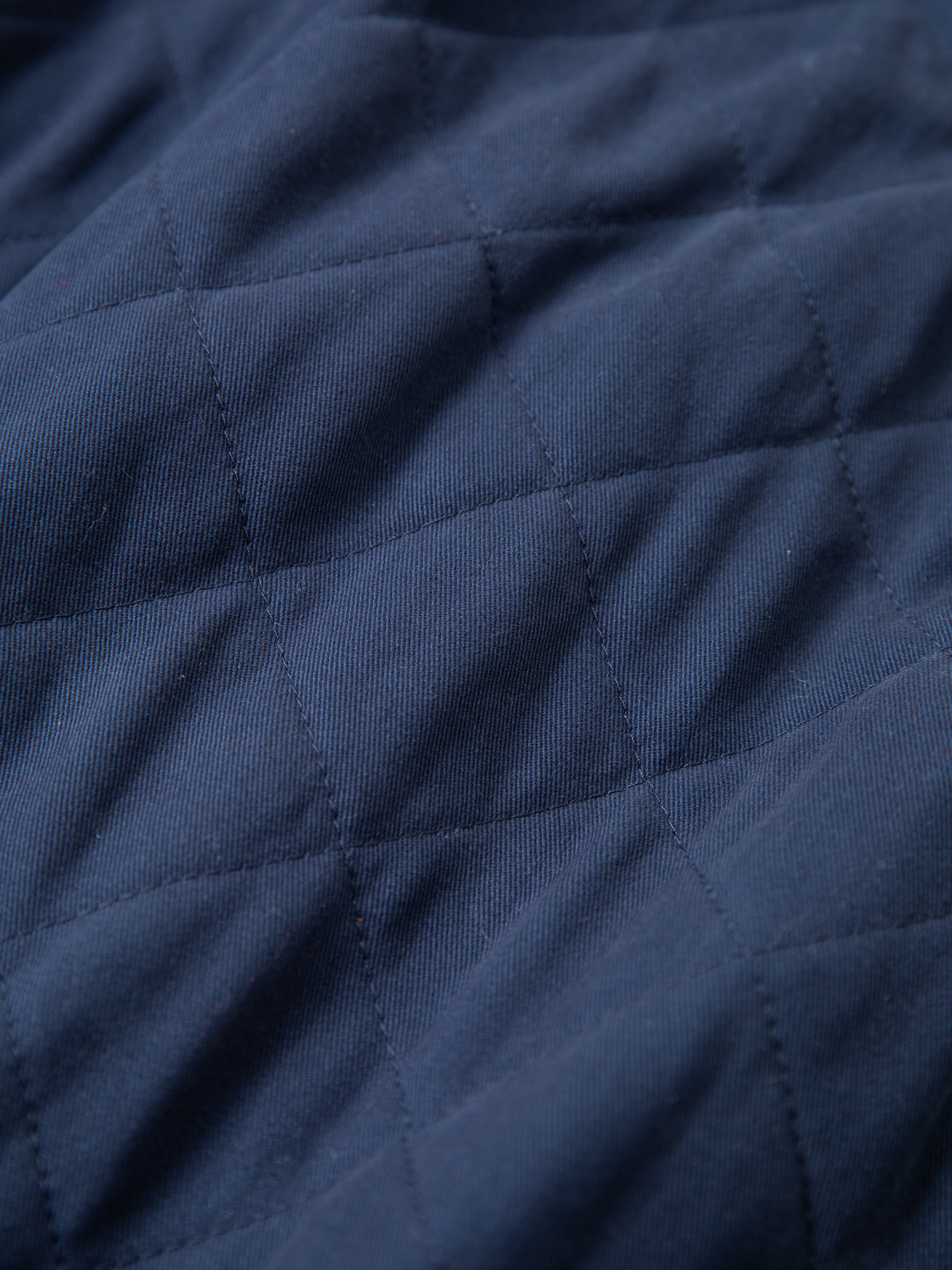 A close-up of a blue, quilted fabric with visible diamond-pattern stitching, reminiscent of the classic Farmstead Quilt Patch Jacket by FOUND. The fabric appears textured and slightly wrinkled, evoking the charm of vintage hunting competition jackets found at farm stands.