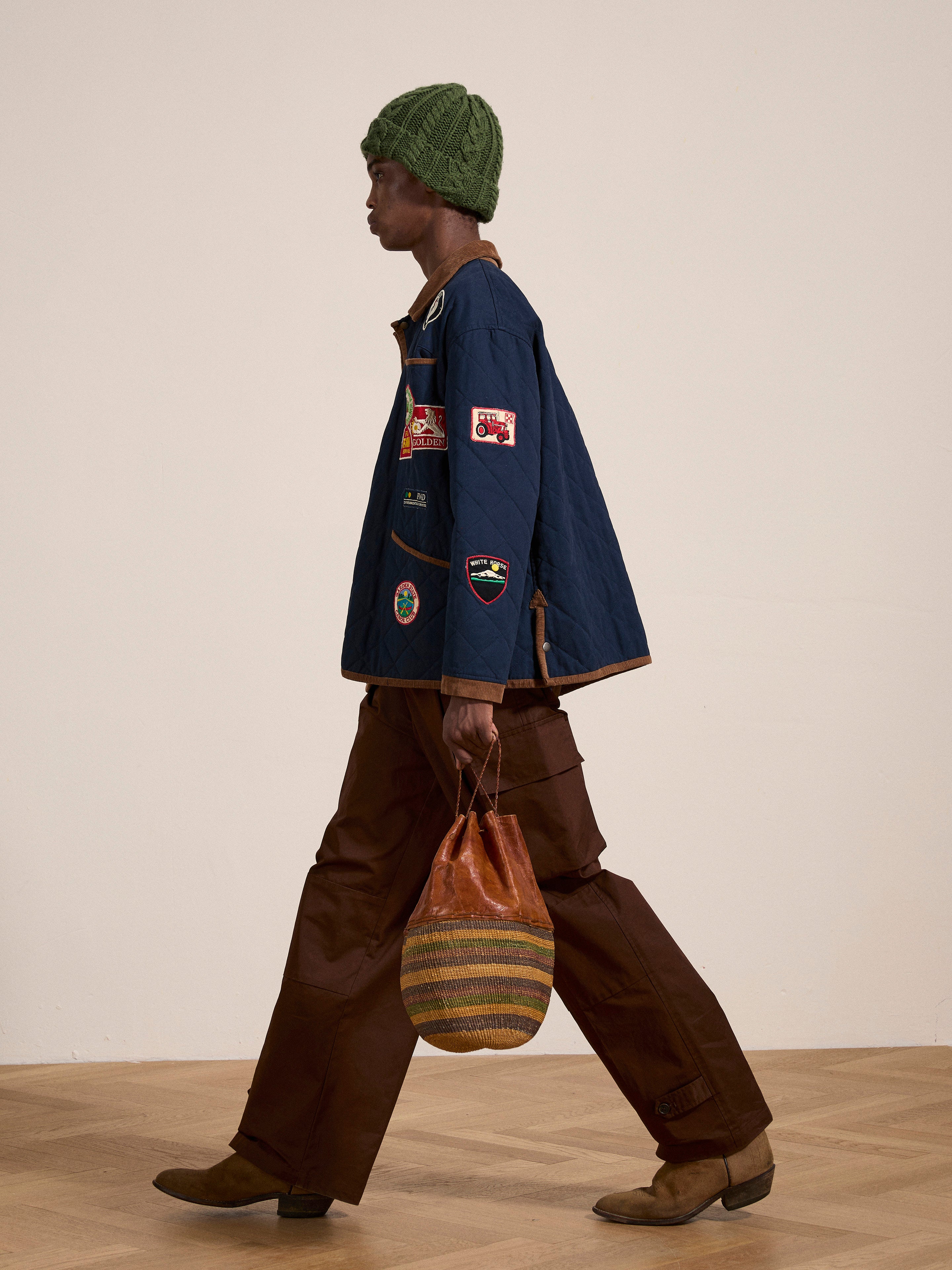 A person is walking while carrying a brown striped bag. They are wearing a green knit hat, a blue Farmstead Quilt Patch Jacket by FOUND, brown pants, and brown boots.