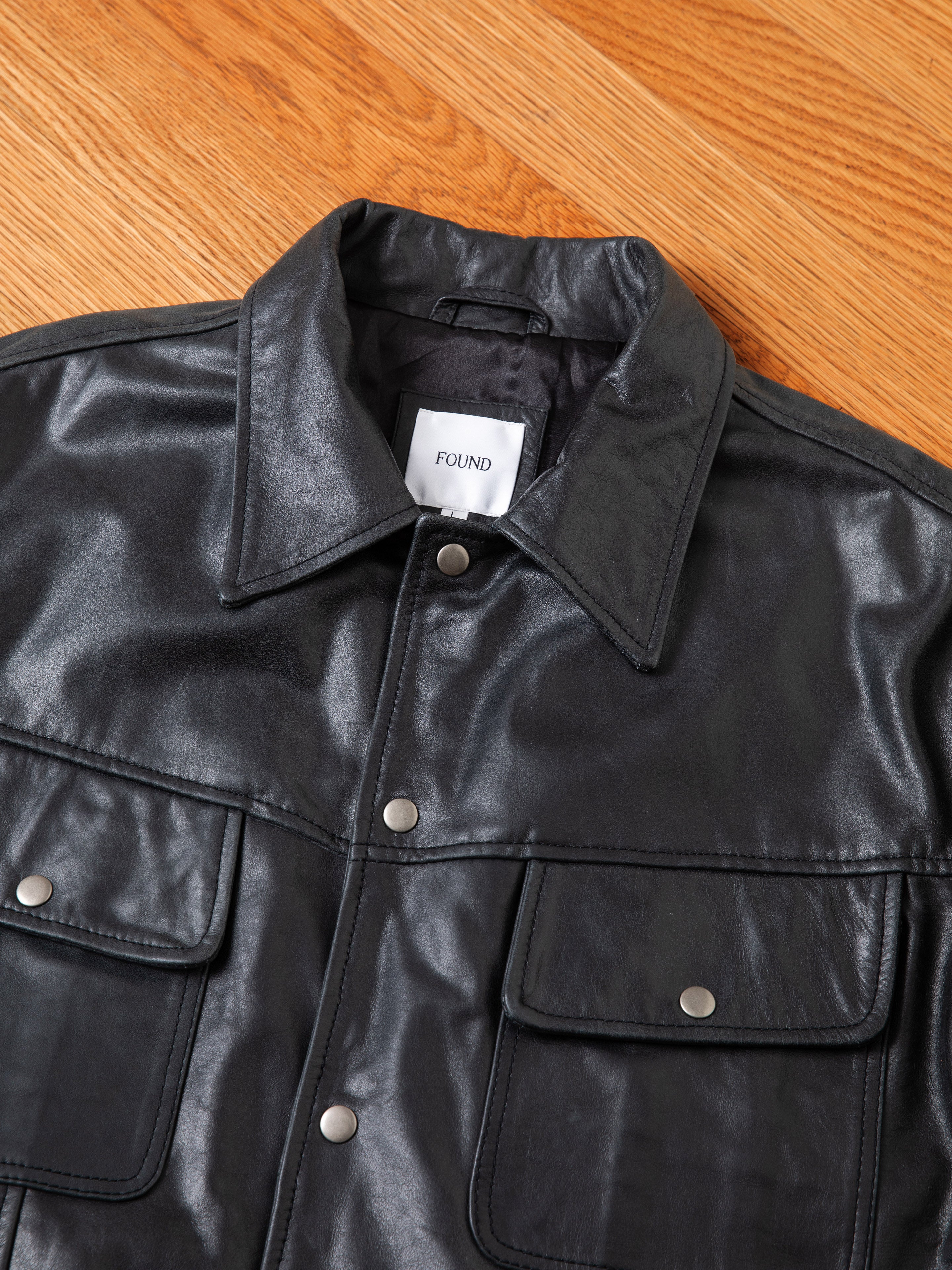 A black Leather Trucker Jacket by FOUND, featuring silver buttons and two front pockets, is laid flat on a wooden floor. Inside the collar, a white label displays the brand name "FOUND.