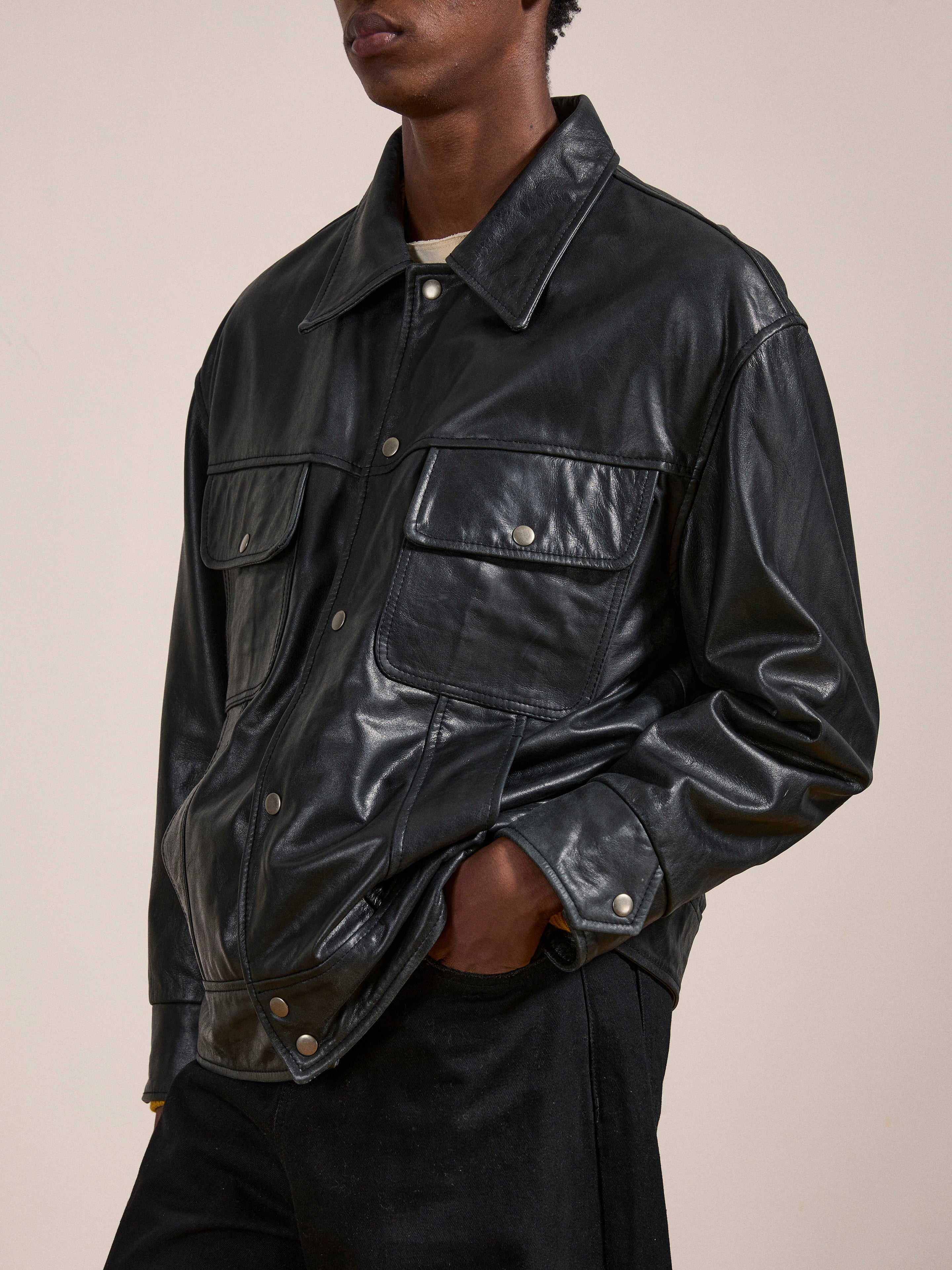 A person is wearing a FOUND Leather Trucker Jacket in classic black, featuring large front pockets and metal buttons. Their hands are tucked into the pockets of their black pants.