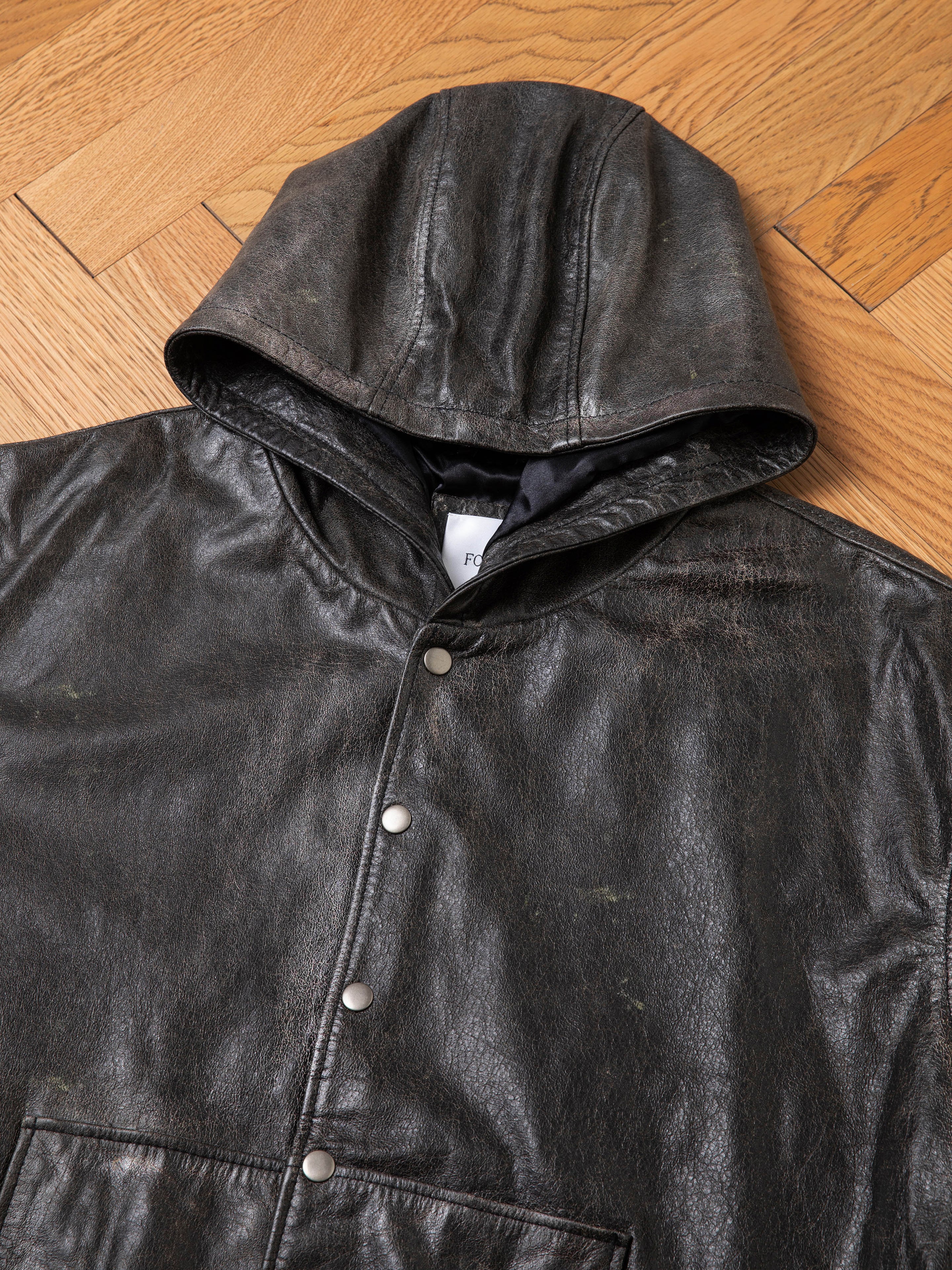 The Found Distressed Genuine Leather Hoodie, featuring metal snap buttons and dark brown distressed leather, is laid flat on a wooden parquet floor.