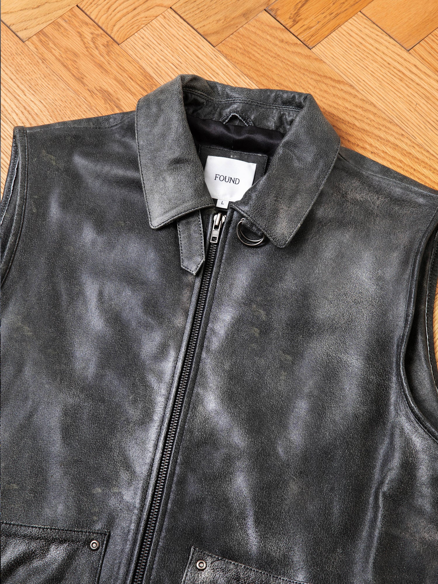 A Distressed Genuine Leather Pocket Vest from Found, featuring a front zipper, is laid flat on a wooden floor. The inside tag reads 