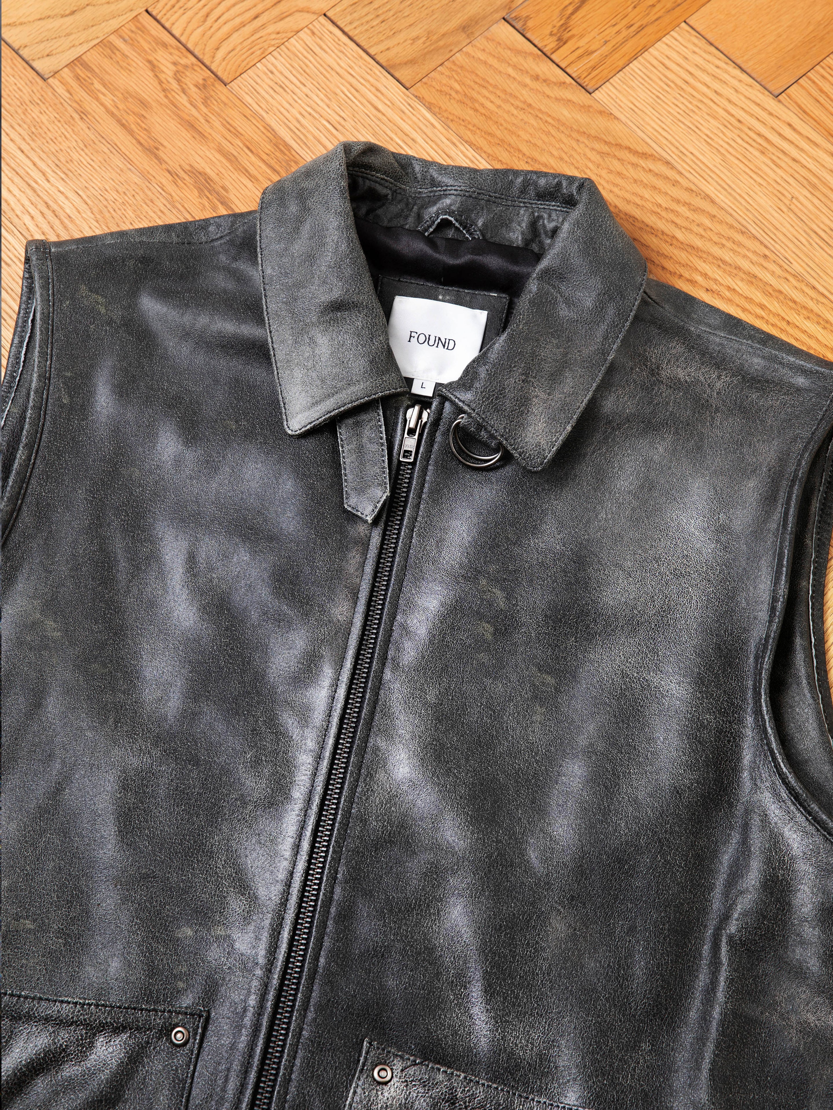 A Distressed Genuine Leather Pocket Vest from Found, featuring a front zipper, is laid flat on a wooden floor. The inside tag reads "Found." Made from ethically sourced materials, this piece combines rugged style with responsible fashion.