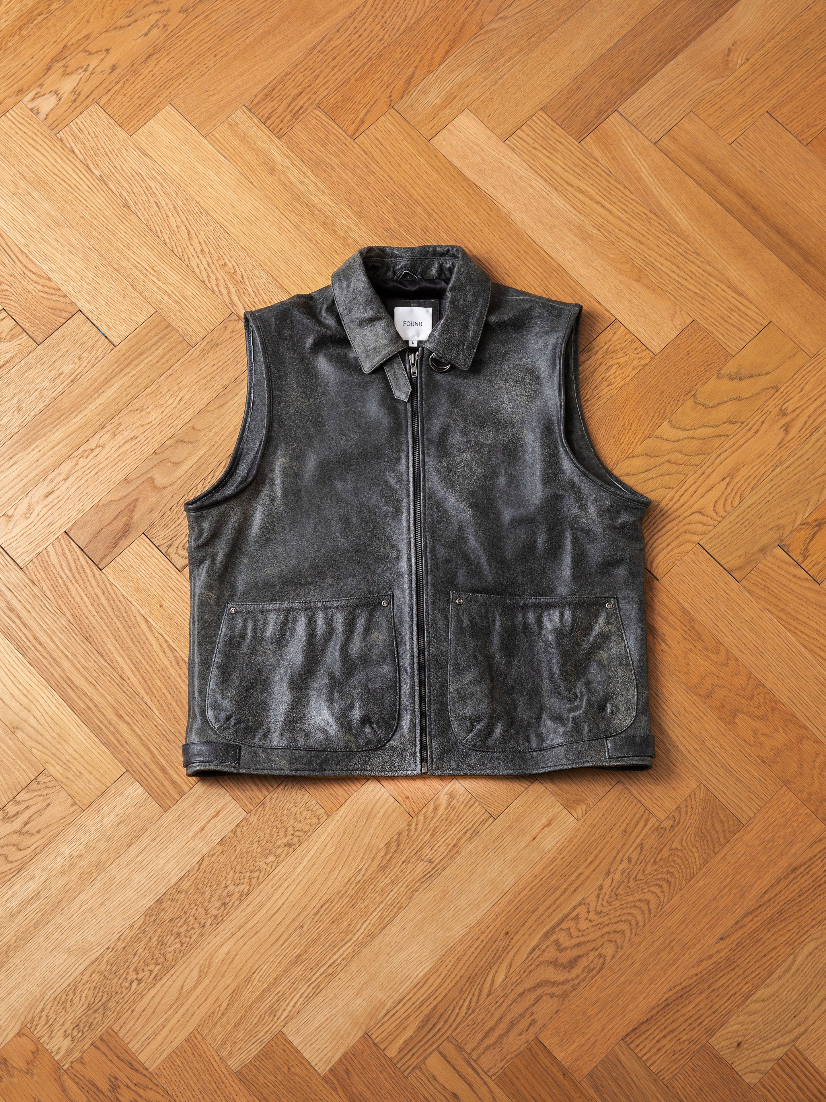 A Distressed Genuine Leather Pocket Vest by Found in dark gray, featuring a zipper closure and two front pockets, is lying flat on a herringbone wooden floor.