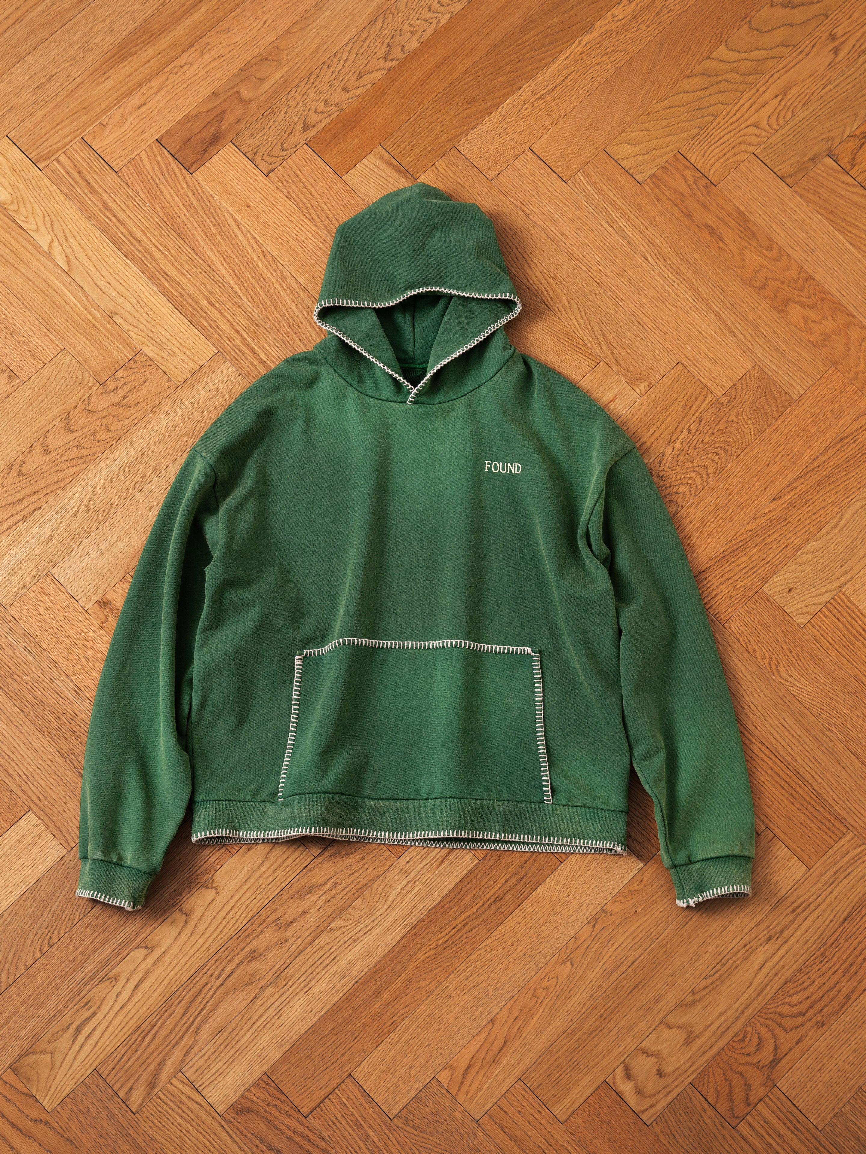 An oversized green hoodie with a front pocket, emblazoned with the "FOUND" logo on the chest, displayed on a wooden herringbone-pattern floor. The Stitched Border Distressed Logo Hoodie offers a vintage feel and added character with its stitched border distressed logo.