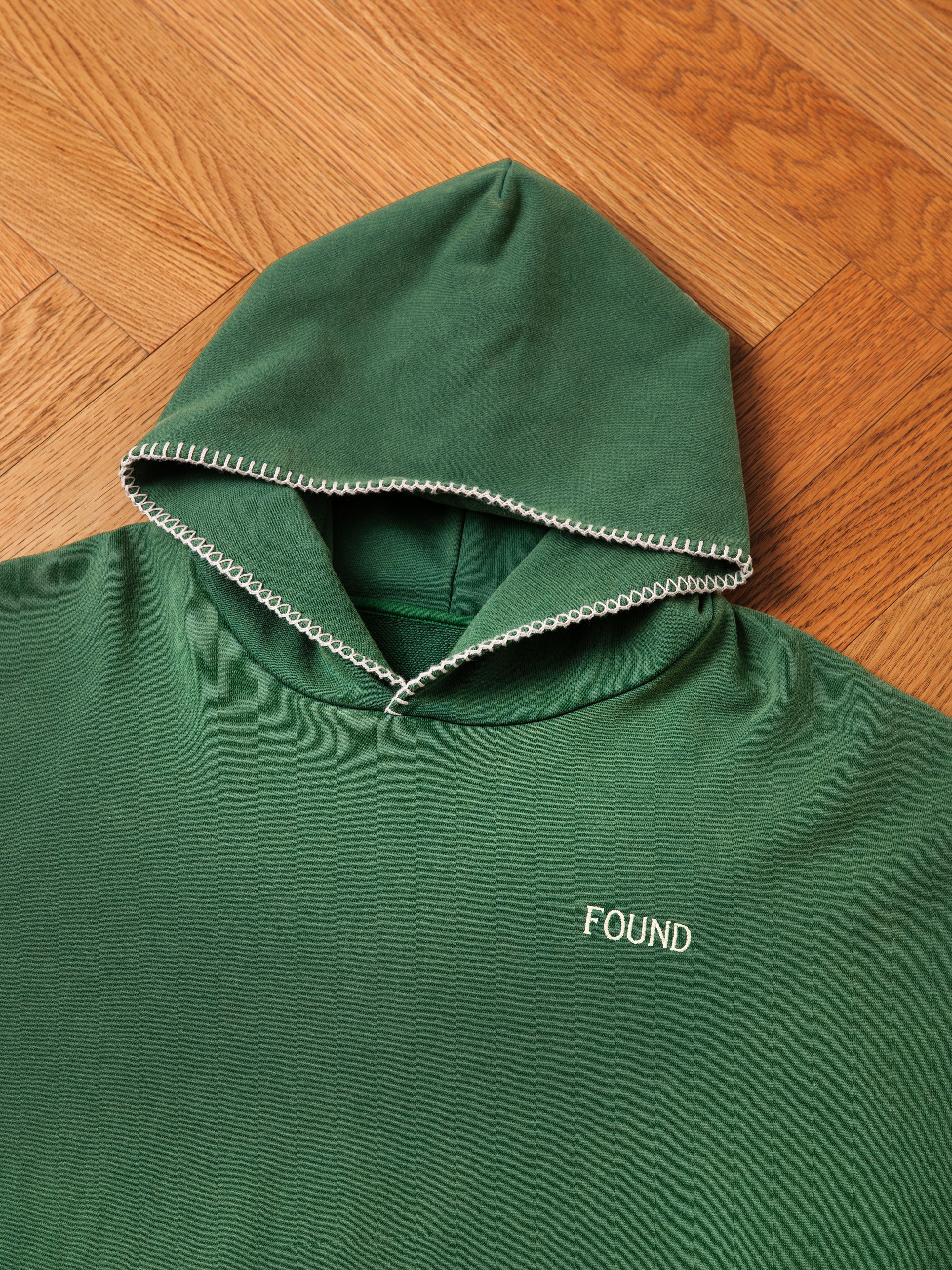 A green, oversized Stitched Border Distressed Logo Hoodie by FOUND, featuring "FOUND" embroidered on the front and a stitched border distressed logo for a vintage feel, is laid flat on a wooden floor.