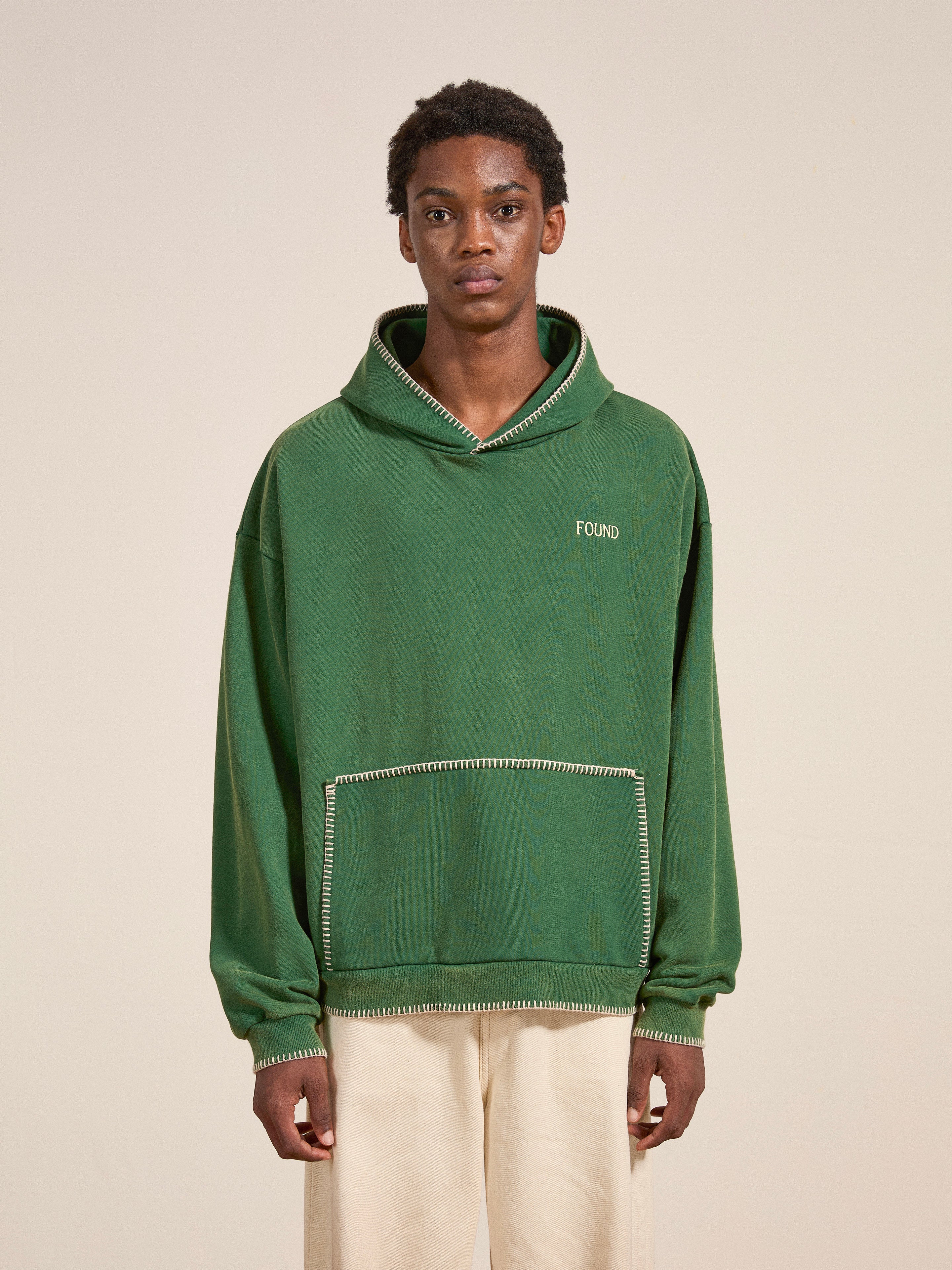 A person stands facing forward, wearing the Stitched Border Distressed Logo Hoodie by FOUND in green and paired with cream-colored pants. The oversized hoodie features a distressed "FOUND" logo and is designed with a stitched border for a vintage feel, contrasting against the plain, light-colored background.