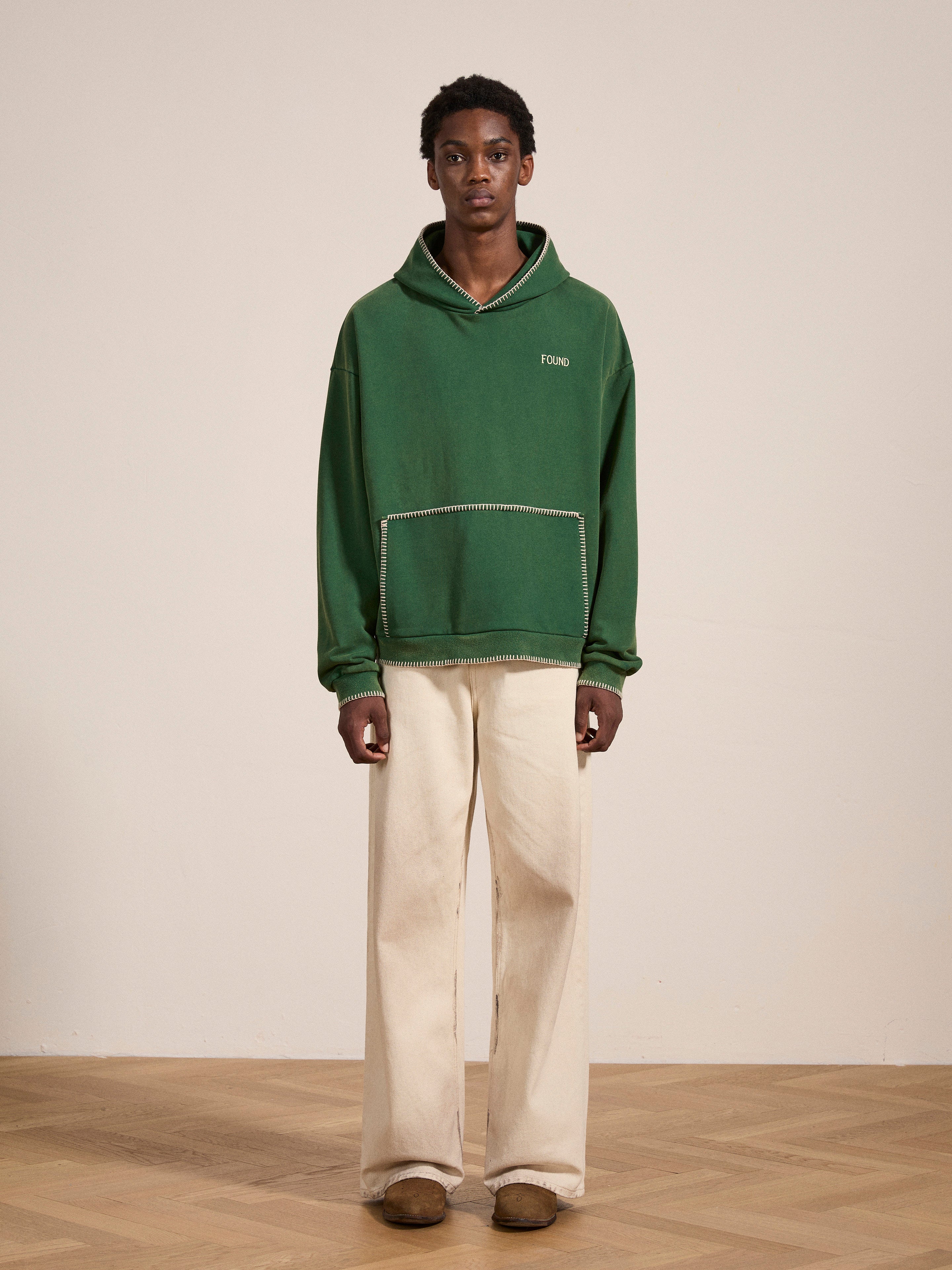 A person stands against a plain backdrop, wearing a green FOUND Stitched Border Distressed Logo Hoodie and light-colored pants. They have a neutral expression, and the floor appears to be wooden, adding to the scene's vintage feel.