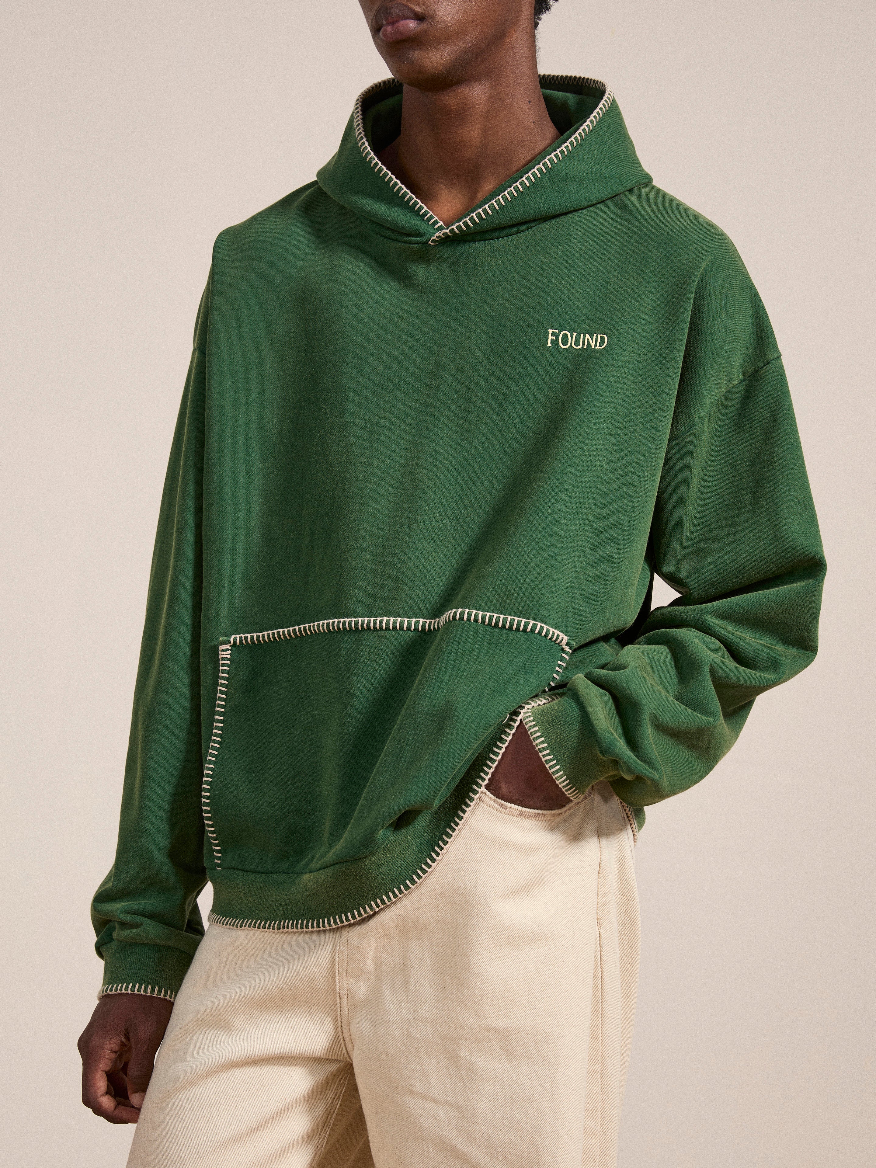 A person is wearing a green, oversized Stitched Border Distressed Logo Hoodie by FOUND, which features "FOUND" embroidered on the chest, contrasting stitching on the hems, and a large front pocket that gives it a vintage feel. They are also wearing beige pants.