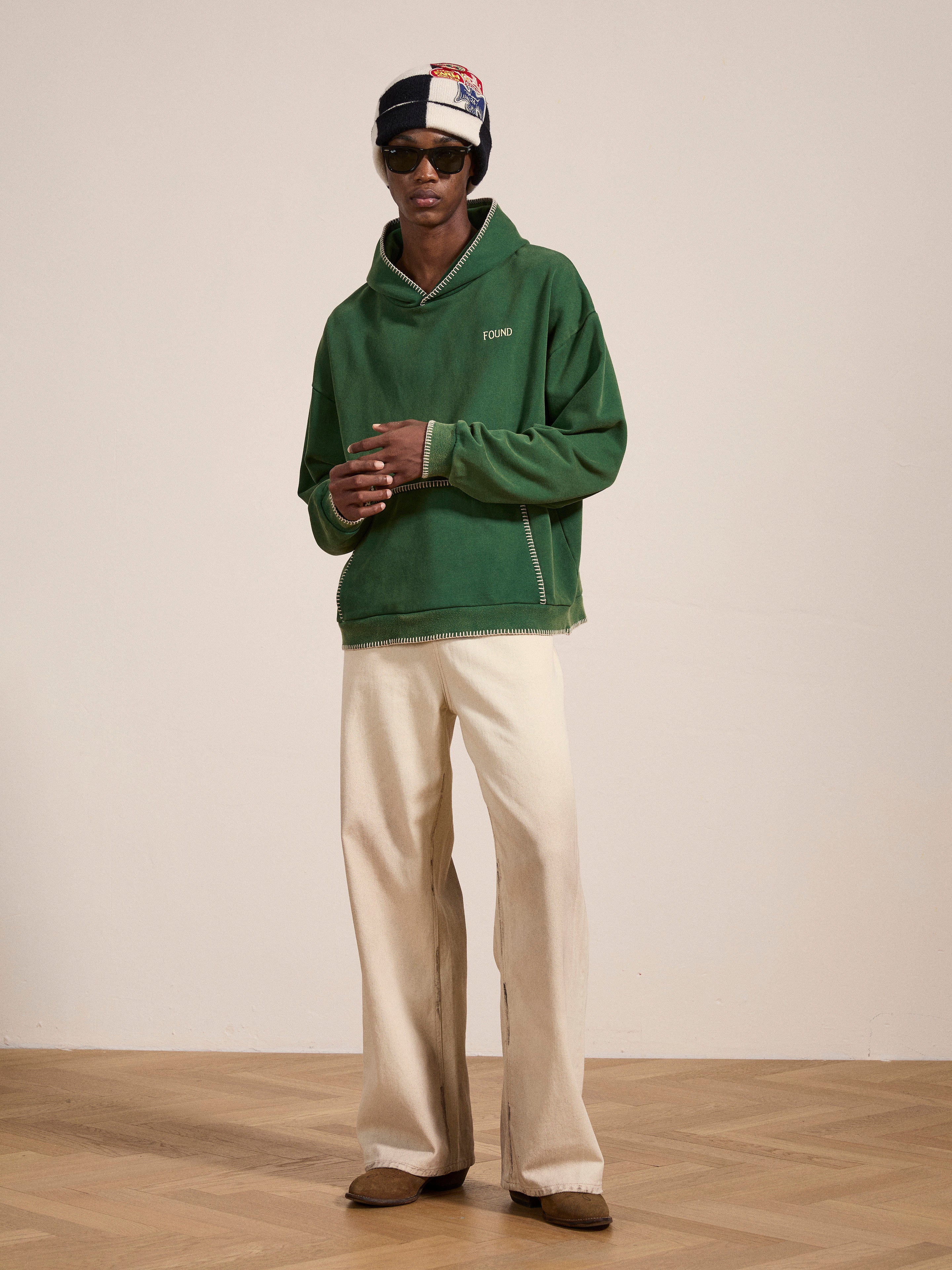 A person wearing the FOUND Stitched Border Distressed Logo Hoodie in an oversized green style, paired with cream wide-leg pants, sunglasses, and a cap stands in a minimalistic indoor setting with a wooden floor. The overall look carries a vintage feel.