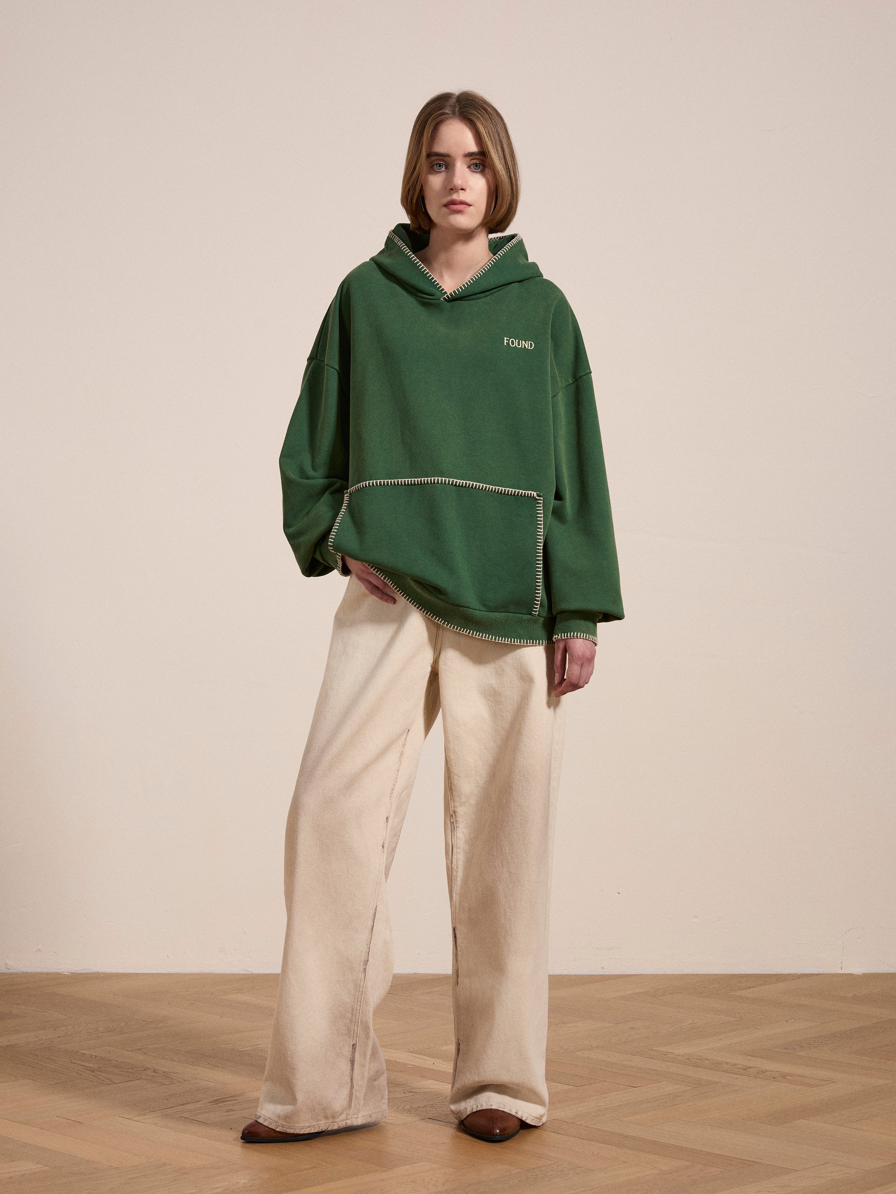 A person sporting a green Stitched Border Distressed Logo Hoodie from FOUND and beige wide-leg pants stands on a wooden floor against a plain background, exuding a vintage vibe.