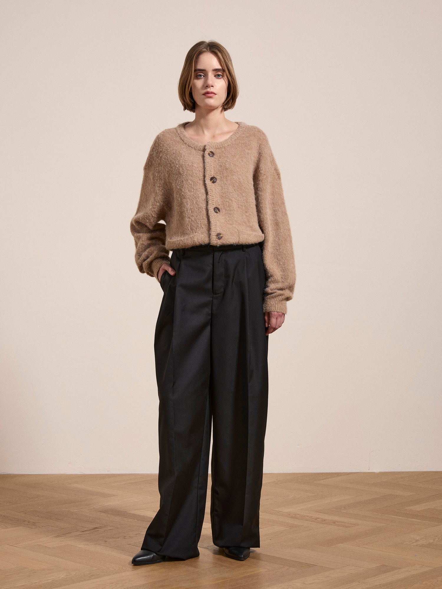 A woman stands indoors on a wooden floor, wearing an oversized FOUND Mohair Cardigan with tortoise buttons and black wide-leg pants. She is looking at the camera with a neutral expression.