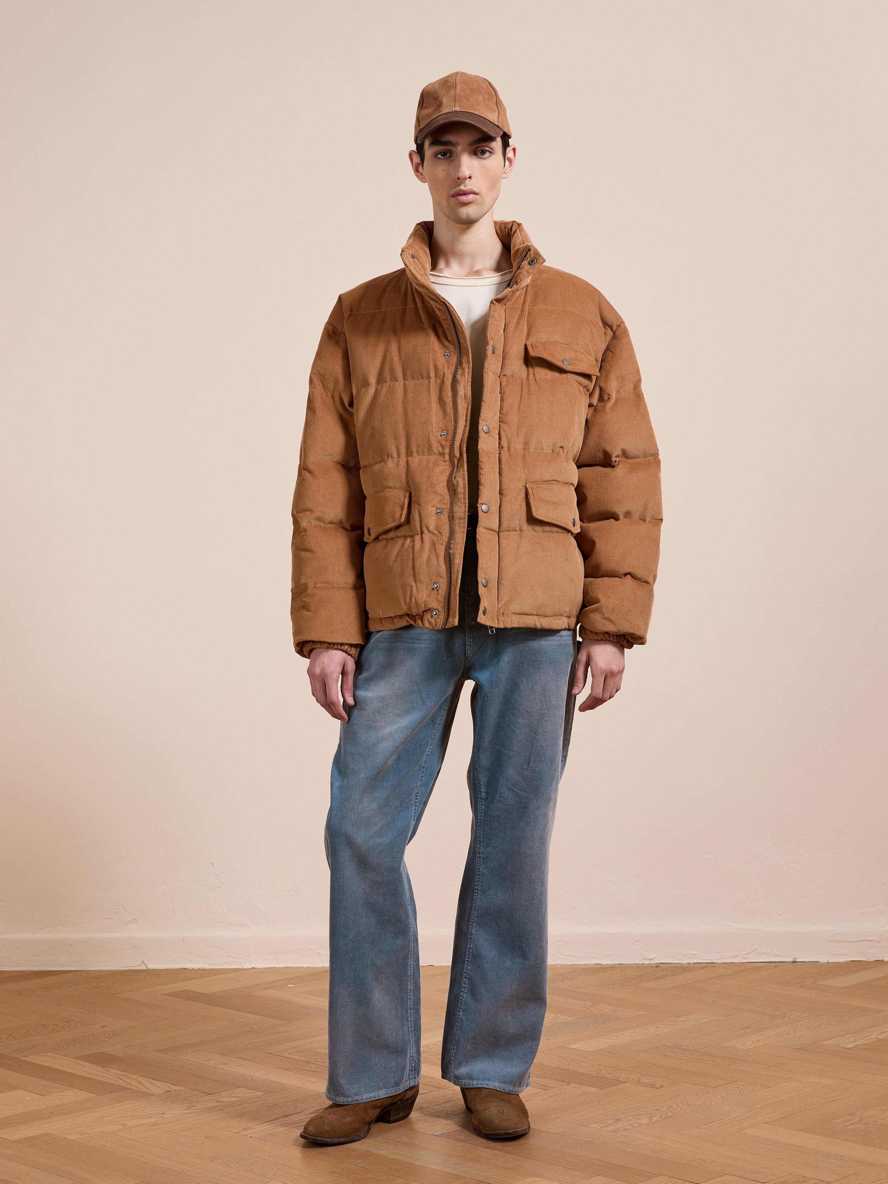 A person stands indoors against a beige wall, facing forward, dressed in a FOUND Corduroy Puffer Jacket, blue jeans, brown shoes, a white shirt, and a brown cap.