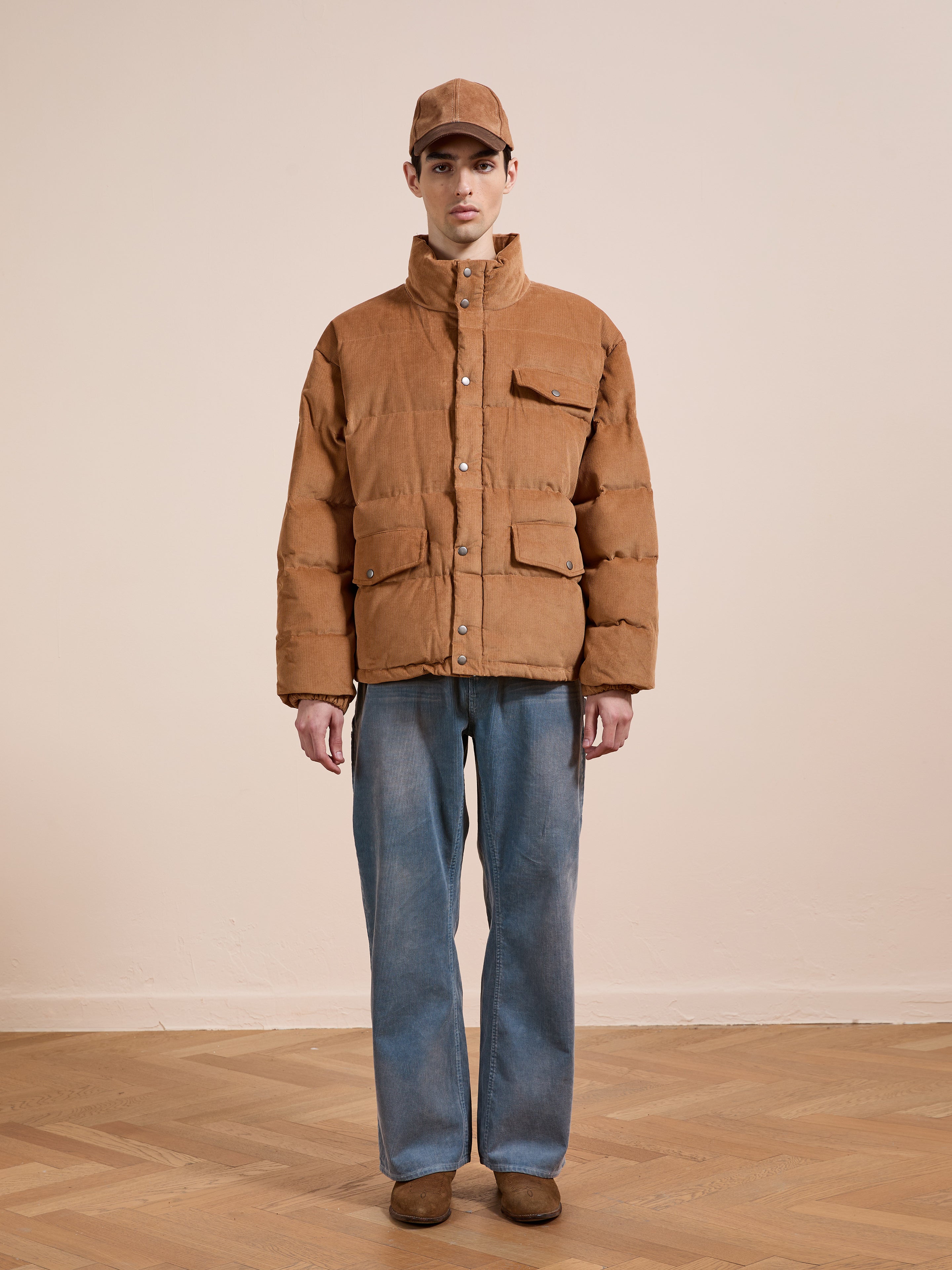 A person stands indoors facing the camera against a plain light wall, wearing the FOUND Corduroy Puffer Jacket in tan, paired with blue jeans, brown shoes, and a matching tan cap.
