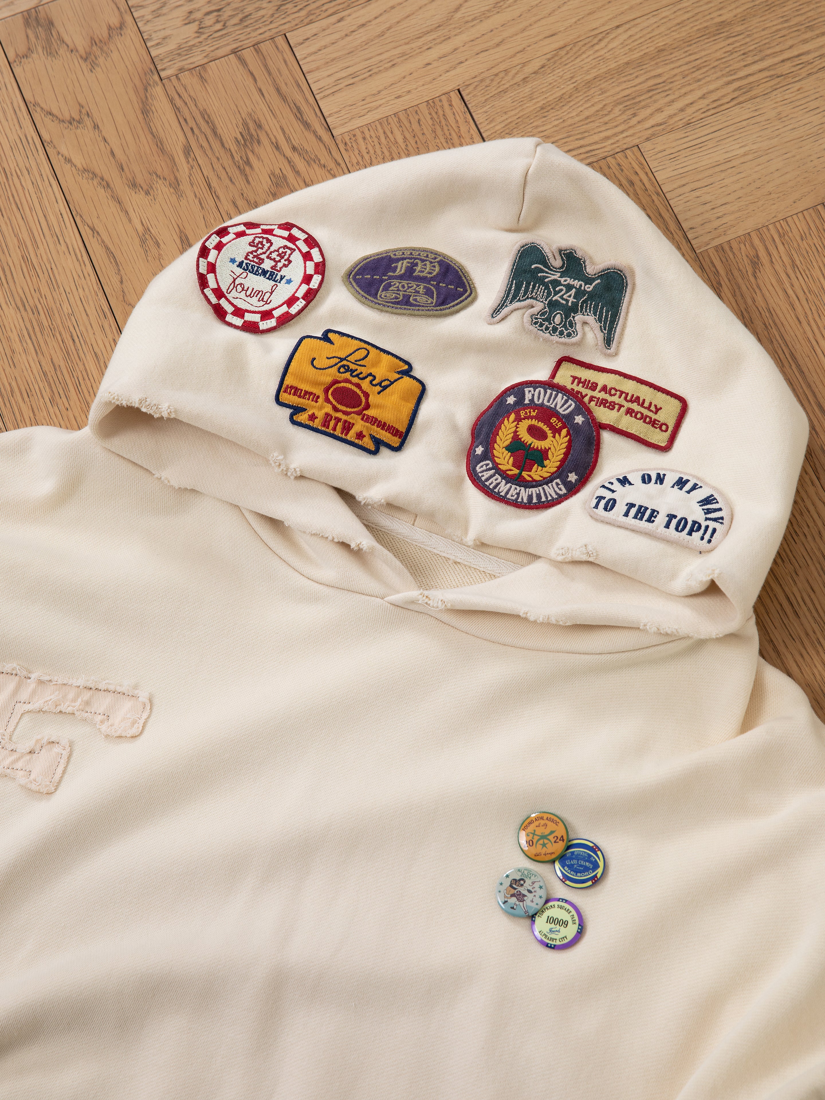 Introducing the Faded Patch Hoodie from FOUND, offering an oversized fit and a vintage aesthetic. This cream-colored hoodie is adorned with a variety of colorful patches on the hood, showcasing unique logos and slogans, making it an ideal addition for infusing retro charm into your casual wardrobe.