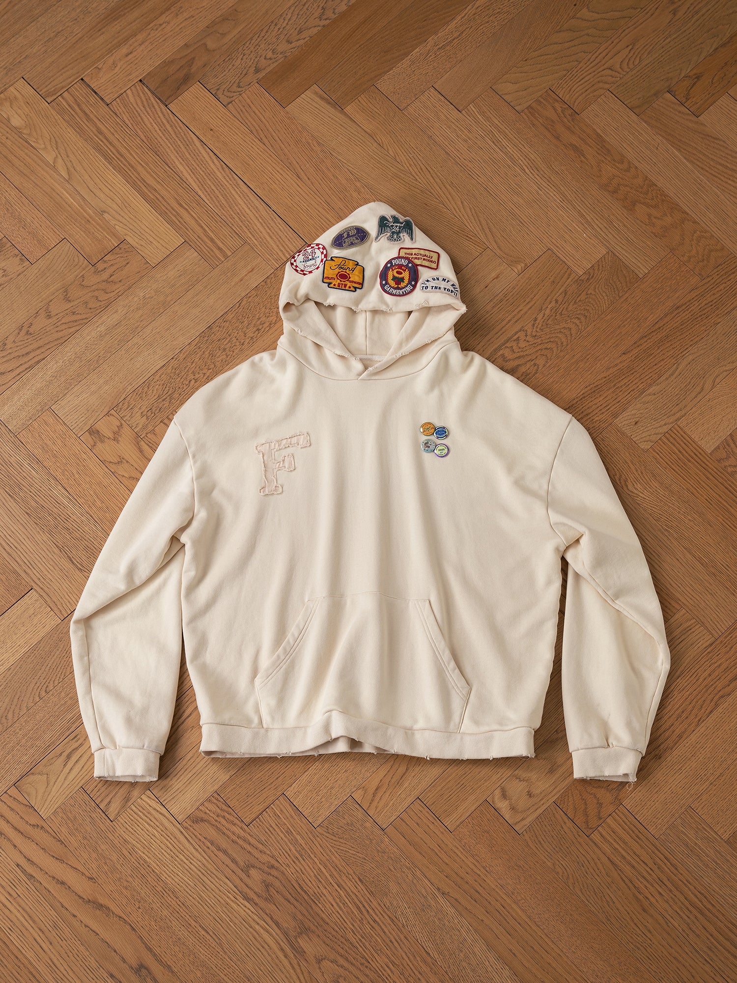 A Faded Patch Hoodie by FOUND, showcasing a vintage cream color adorned with vibrant patches on the hood and chest, is laid flat on a wooden herringbone floor.