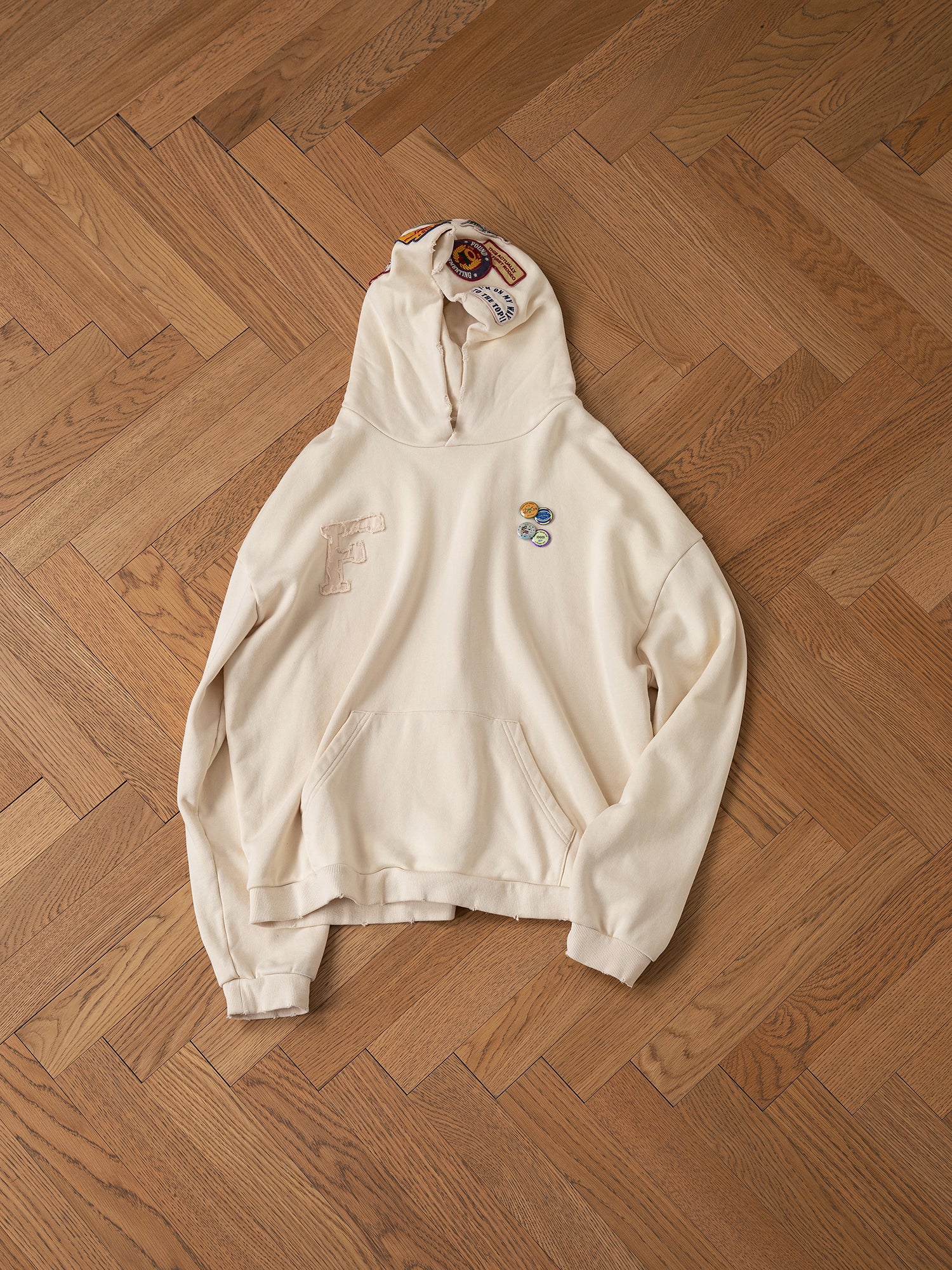 A vintage-inspired Faded Patch Hoodie by FOUND, featuring a cream color, oversized fit, and embroidered patches including an "F" and various symbols, laid flat on a wooden herringbone floor.