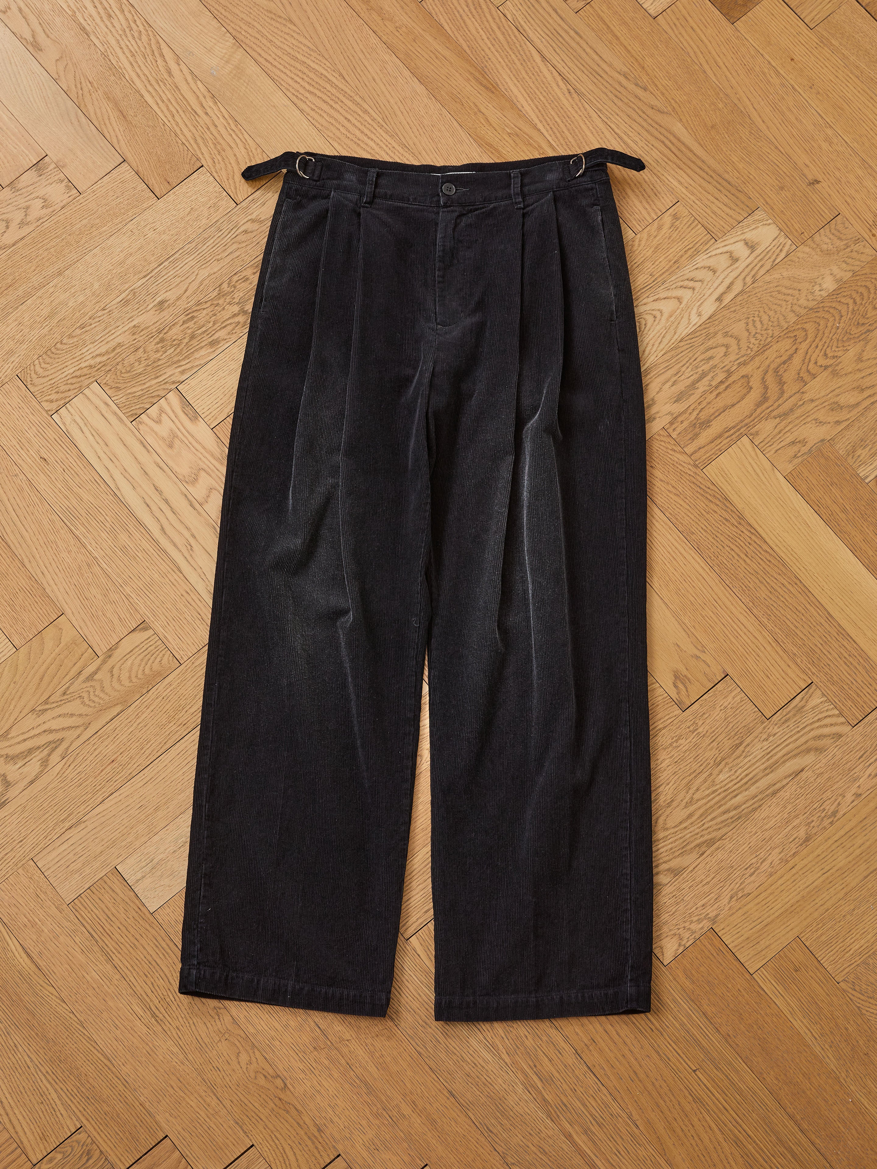 FOUND Faded Pleated Corduroy Pants with wide legs and side adjuster tabs, shown laid flat on a light wood herringbone floor.