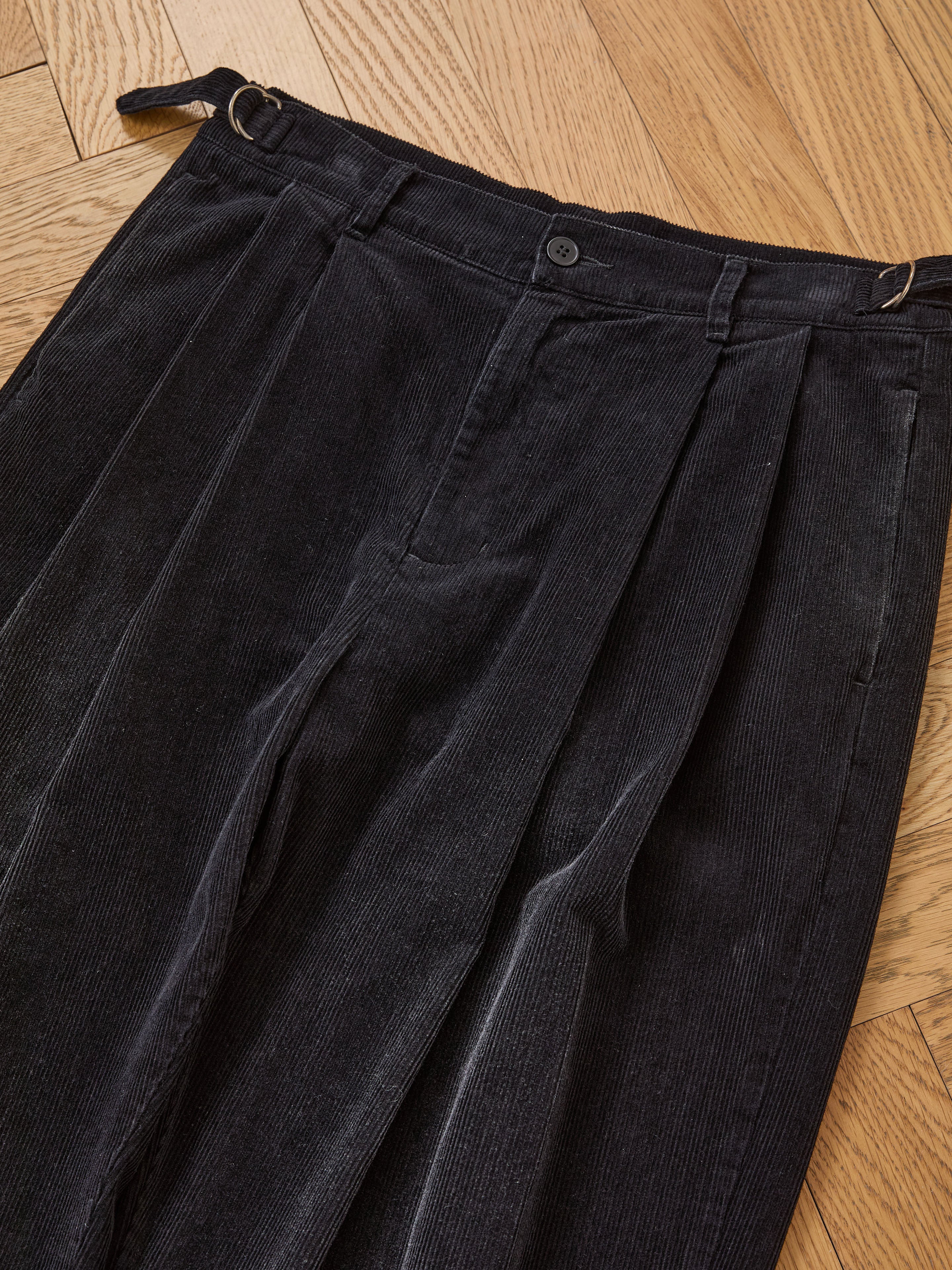 The FOUND Faded Pleated Corduroy Pants, featuring double pleats, a button closure, and adjustable side tabs in black, are shown laid flat on a wooden floor.