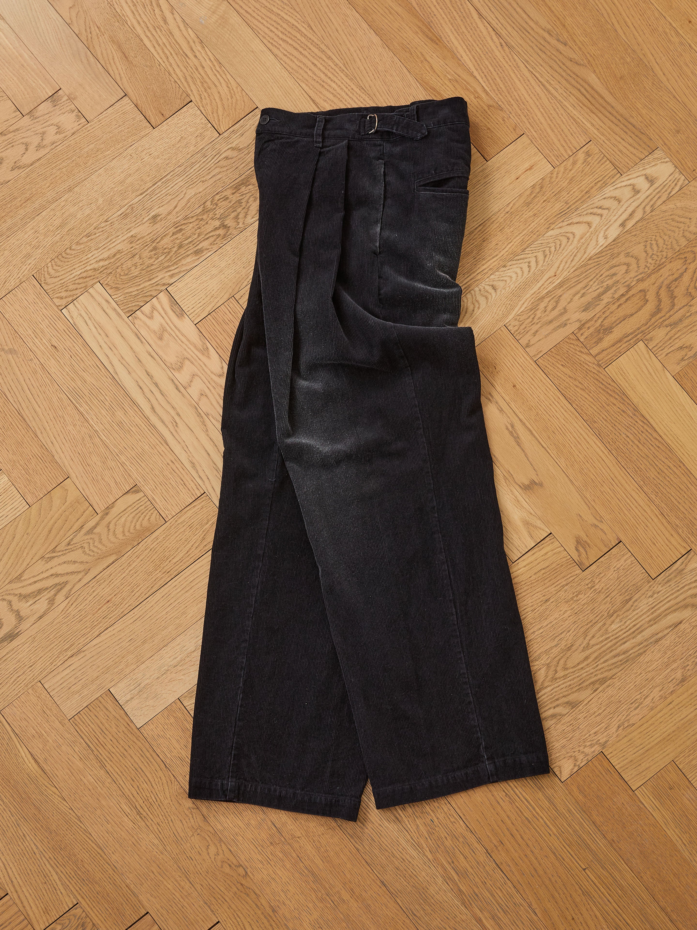 A pair of FOUND Faded Pleated Corduroy Pants is displayed flat on a light wooden herringbone floor.