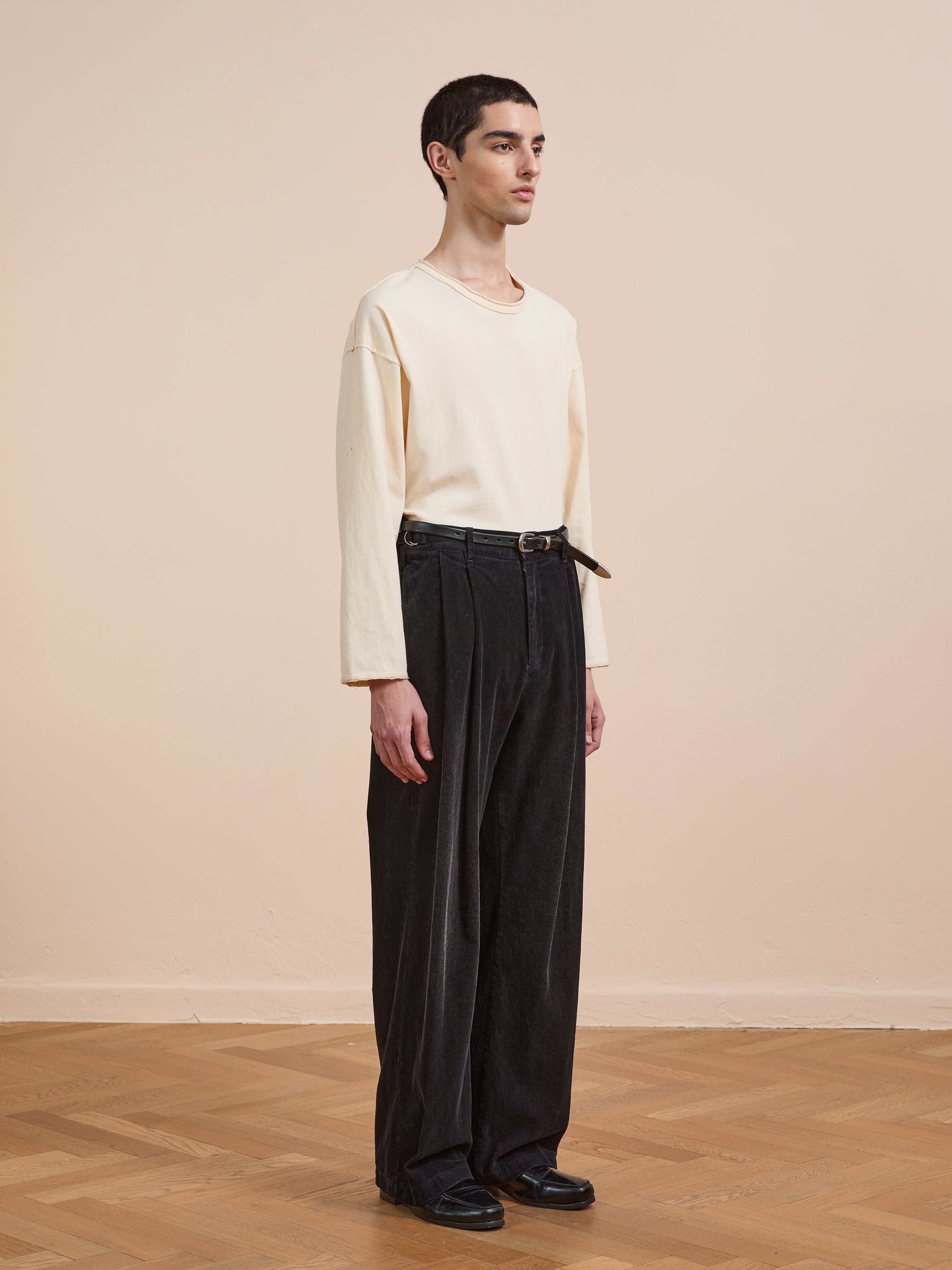 A person stands indoors facing slightly right against a beige wall, wearing a long-sleeve cream shirt, black shoes, and FOUND Faded Pleated Corduroy Pants.