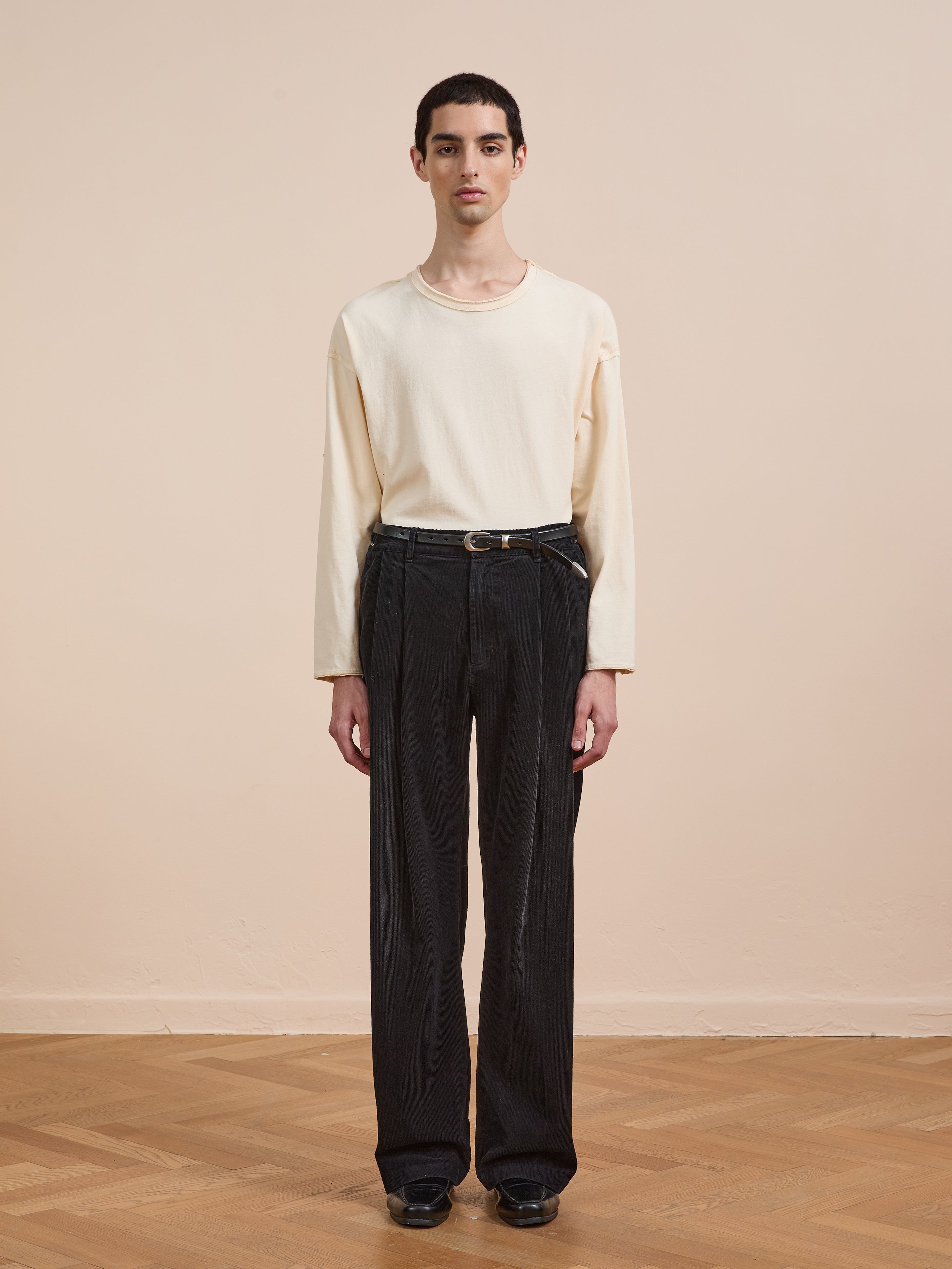 A person stands indoors on a wooden floor, facing the camera against a beige background, wearing a long-sleeve cream shirt and FOUND Faded Pleated Corduroy Pants, styled with a belt and black shoes.