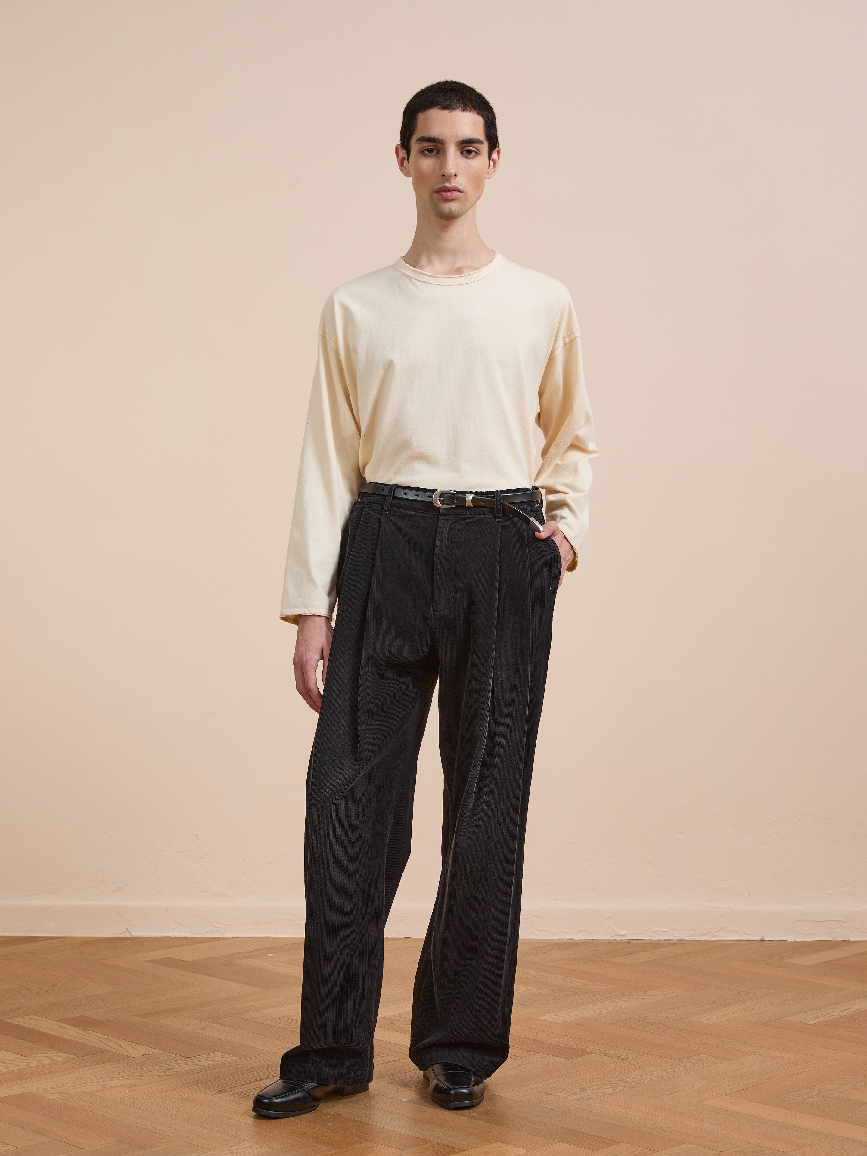 A person stands indoors on a wooden floor, wearing FOUND’s Faded Pleated Corduroy Pants paired with a long-sleeved cream shirt, black shoes, and a belt, set against a plain beige background.