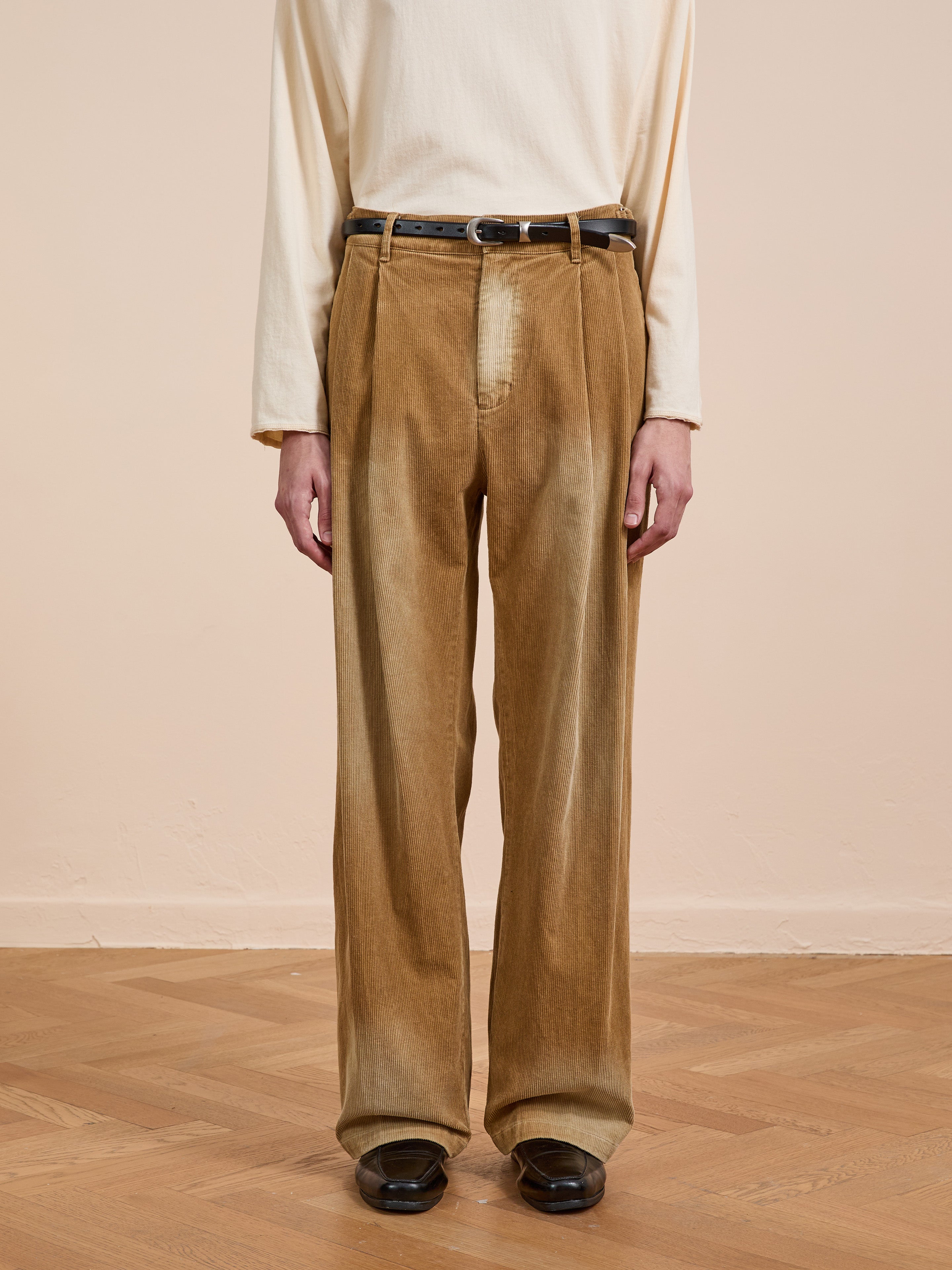 A person wears a cream long-sleeve shirt tucked into FOUND’s Faded Pleated Corduroy Pants with a black belt, standing on a wooden floor against a beige wall.