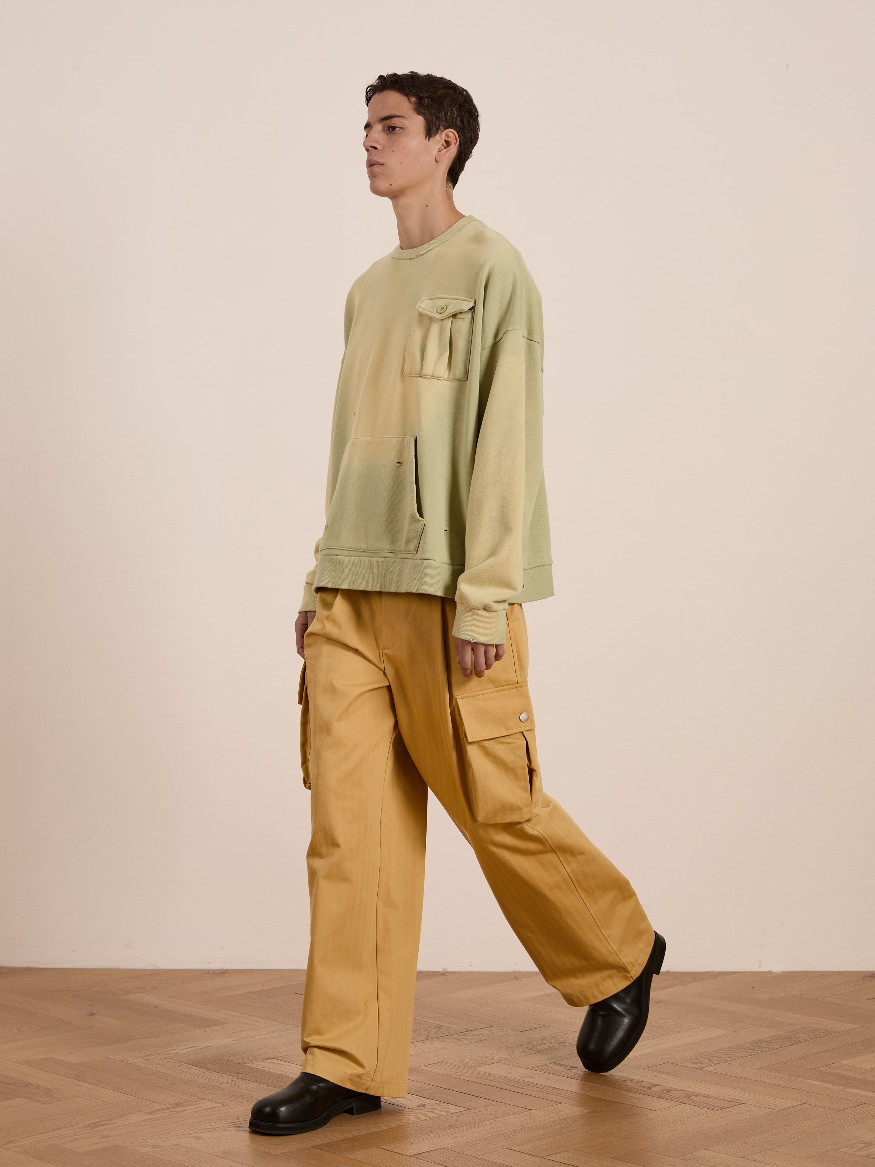 Strolling indoors, a person wears FOUND's Faded Pocket Crewneck, a vintage-inspired green sweatshirt with pocket details. This unisex relaxed fit pairs effortlessly with yellow cargo pants and black shoes for a timeless look suitable for any occasion.