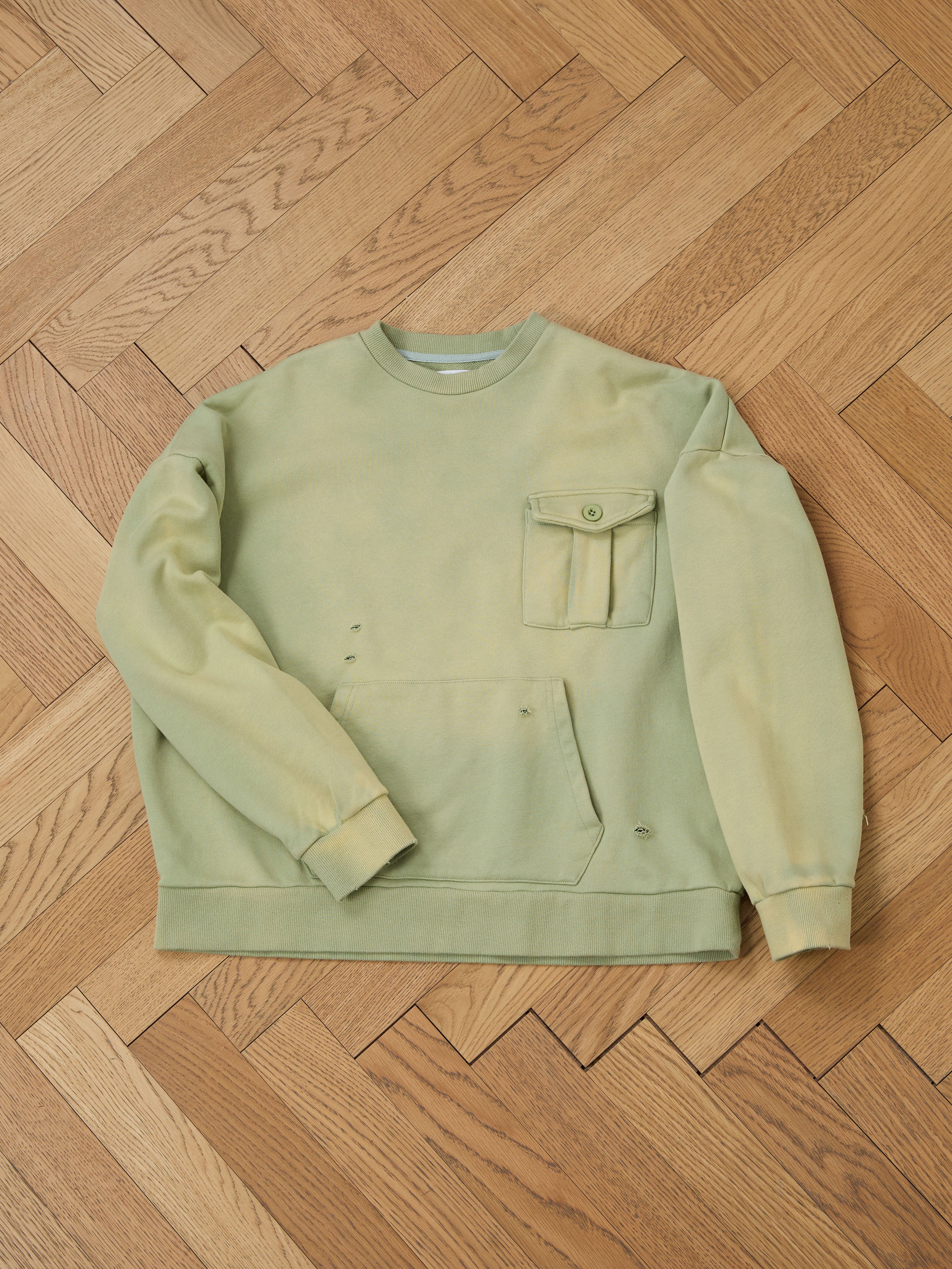 The Faded Pocket Crewneck from FOUND, a vintage-inspired green sweatshirt with a pouch and buttoned chest pocket, is displayed on a wooden herringbone floor. Crafted from heavyweight cotton, it features a unisex relaxed fit.