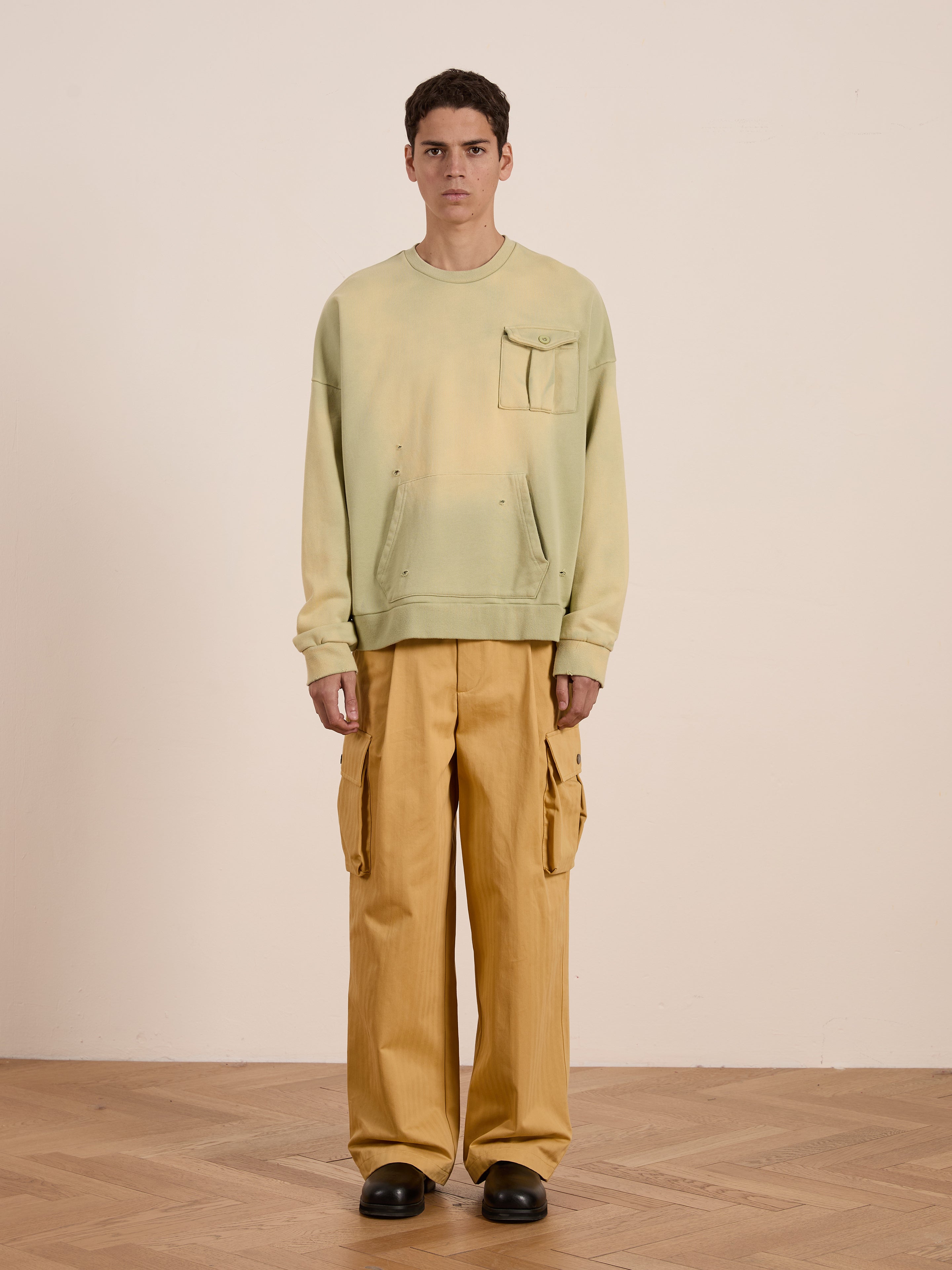 A person stands on a wooden floor against a plain wall, wearing FOUND's vintage-inspired Faded Pocket Crewneck in light green and yellow cargo pants in a unisex relaxed fit.