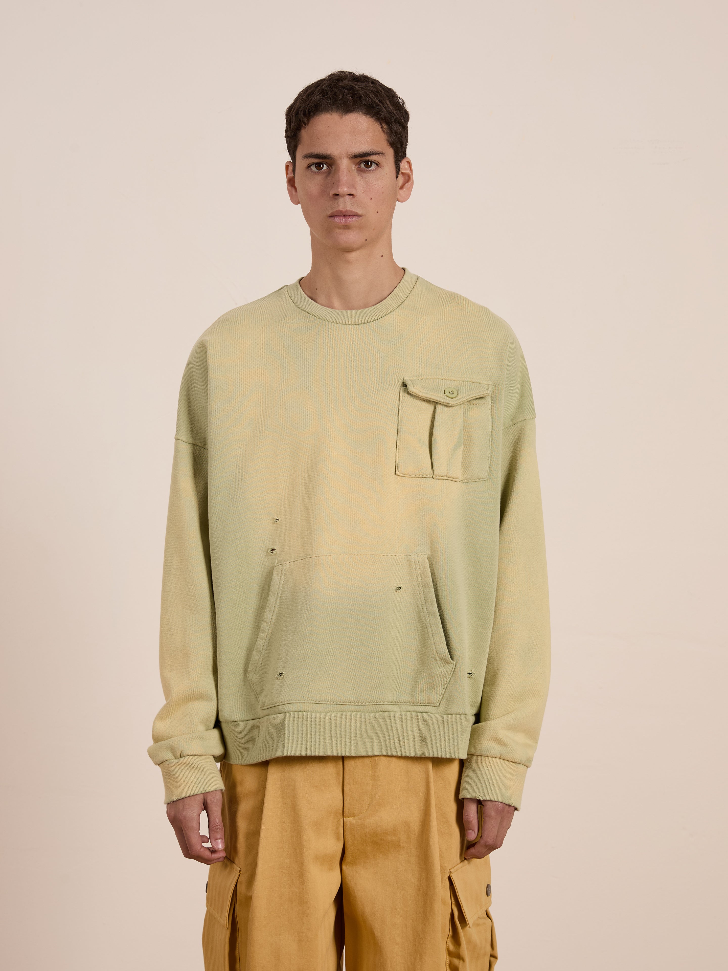 A person wears FOUND's Faded Pocket Crewneck, a light green sweatshirt with a vintage-inspired chest pocket in heavyweight cotton, paired with khaki pants. This unisex relaxed-fit outfit blends comfort and style against a plain backdrop.