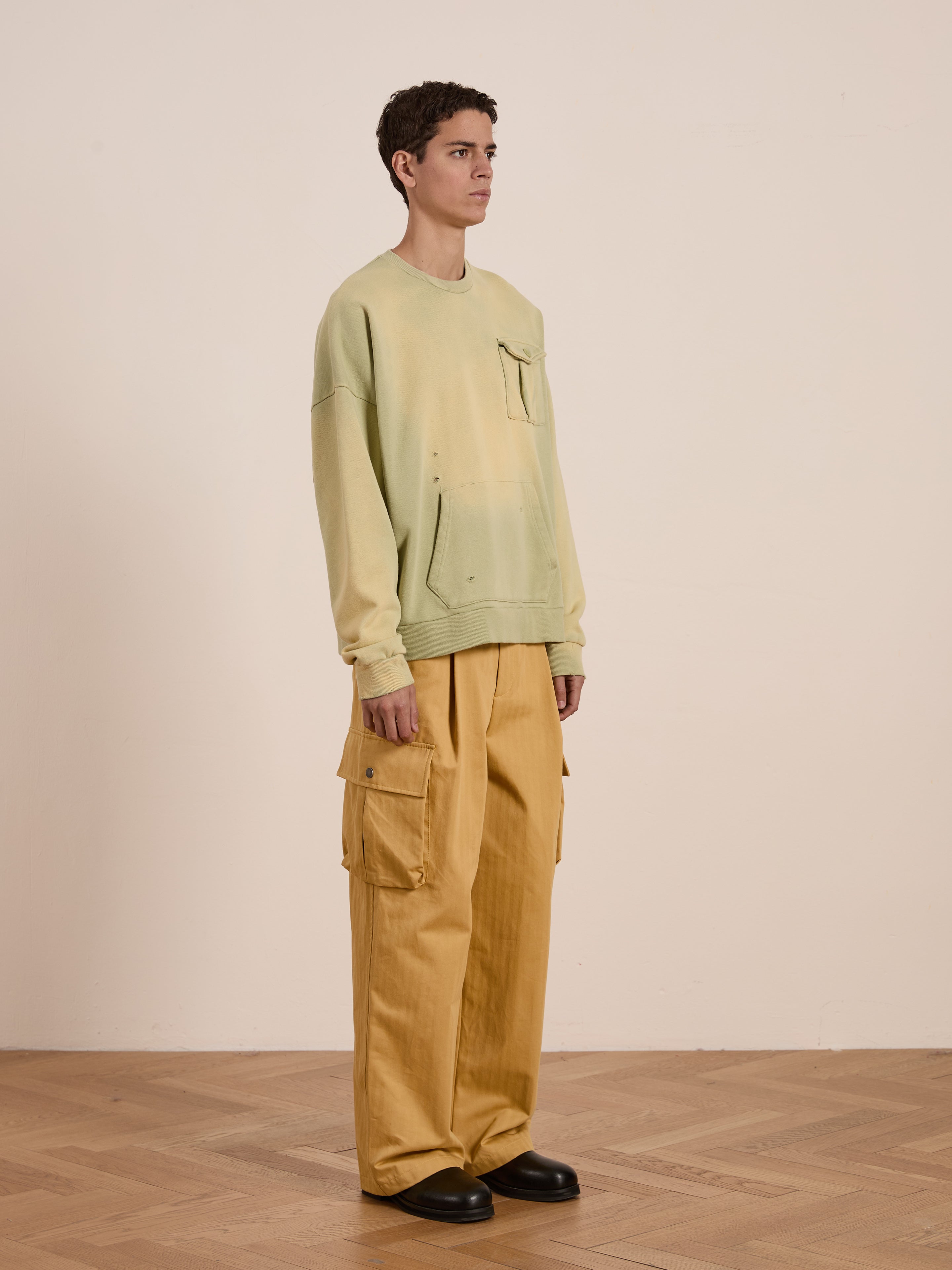 Standing on a wooden floor, a person wears FOUND's Faded Pocket Crewneck, vintage-inspired in light green heavyweight cotton with a front pocket, paired with yellow cargo pants. The unisex ensemble offers a relaxed fit against a plain backdrop.