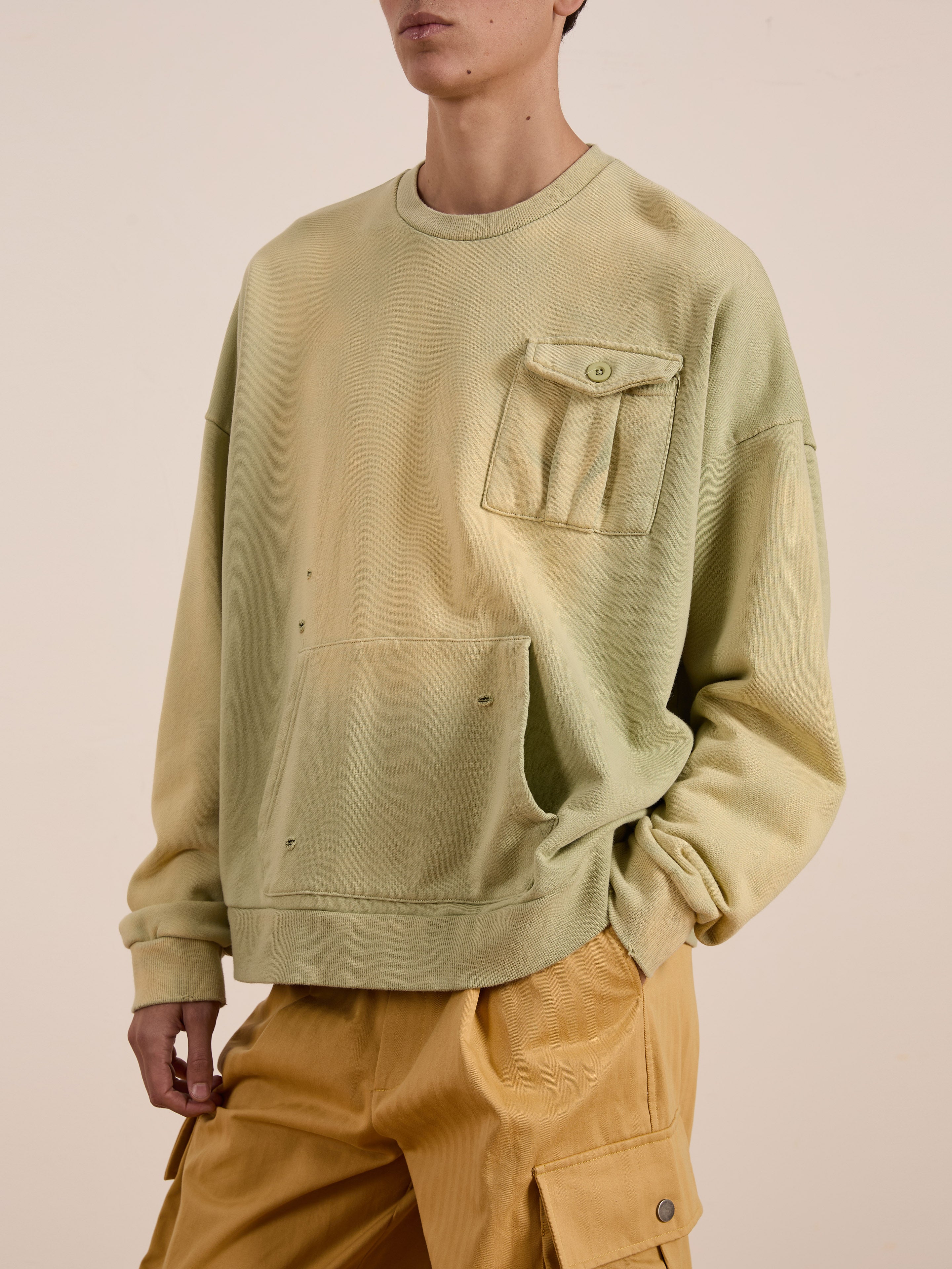 A person wearing the FOUND Faded Pocket Crewneck in a vintage-inspired light olive-green, heavyweight cotton with a chest pocket, and matching unisex relaxed fit shorts stands against a neutral background.