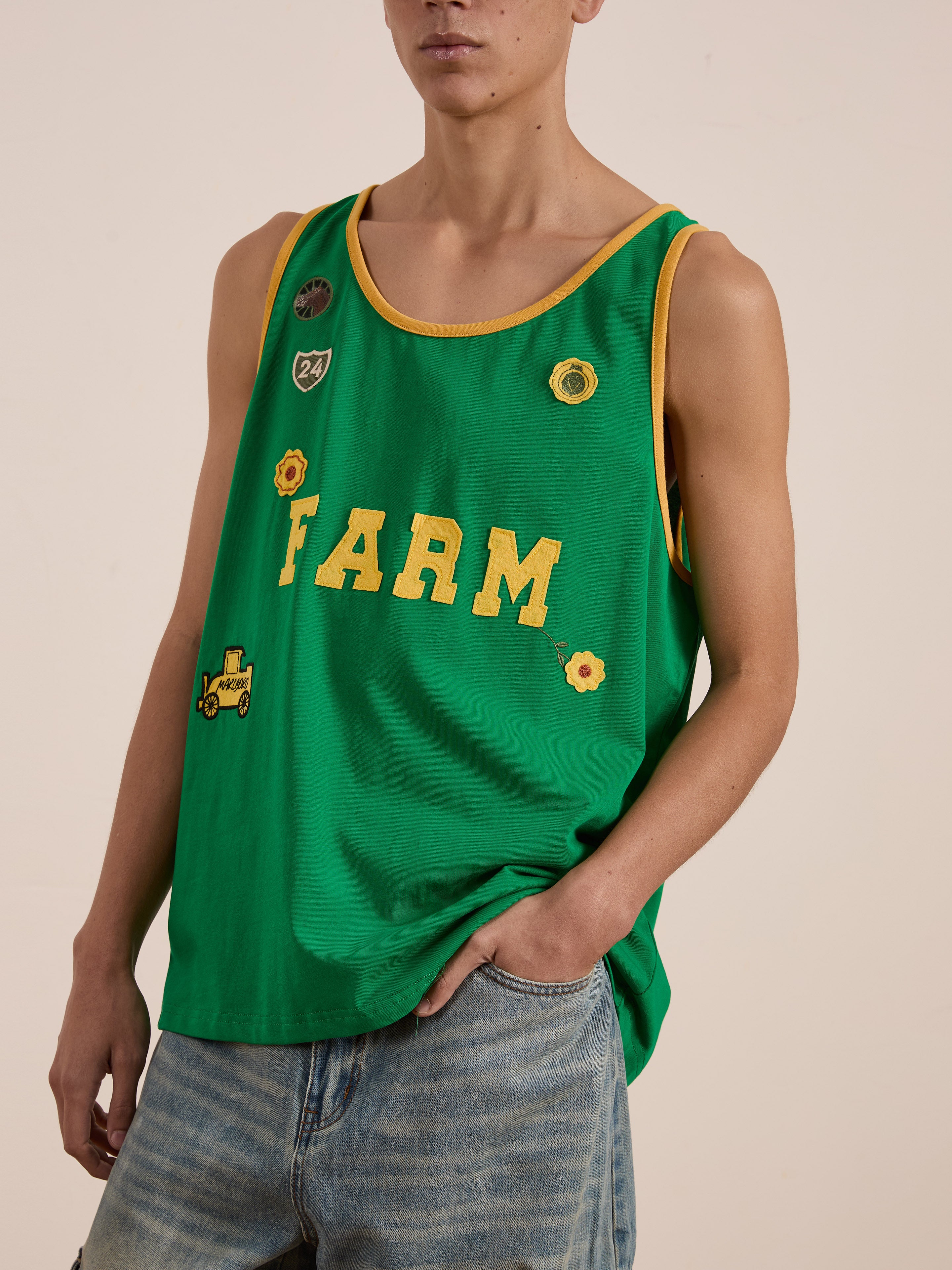 Wearing FOUND's "Farm Tank," a green cotton sleeveless shirt with yellow "FARM" text and embroidered patches, one effortlessly pairs it with blue jeans.