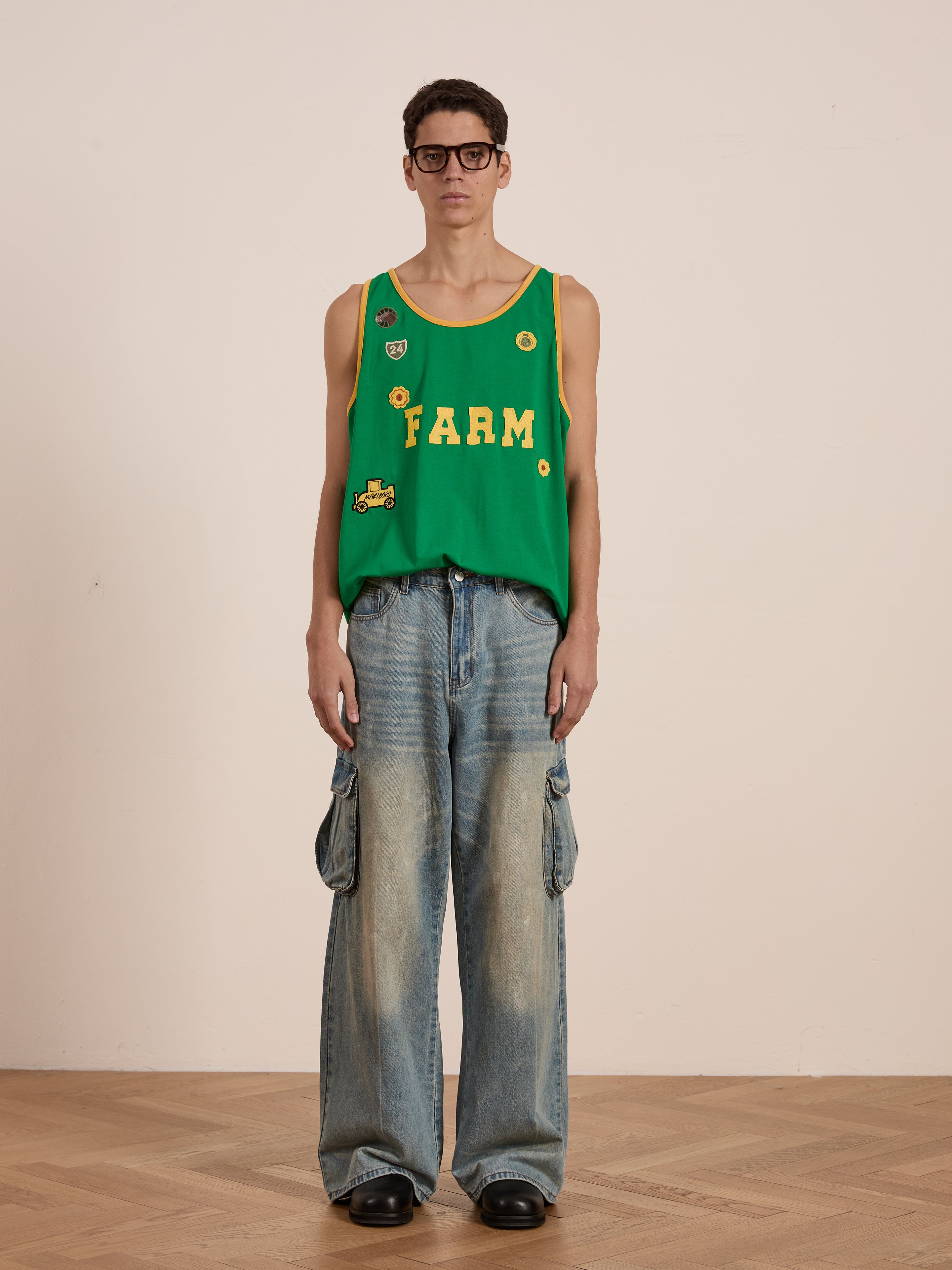 A person in a FOUND "Farm Tank" in green and wide-leg jeans, giving off a vintage athletic vibe, stands on a wooden floor against a plain beige background.