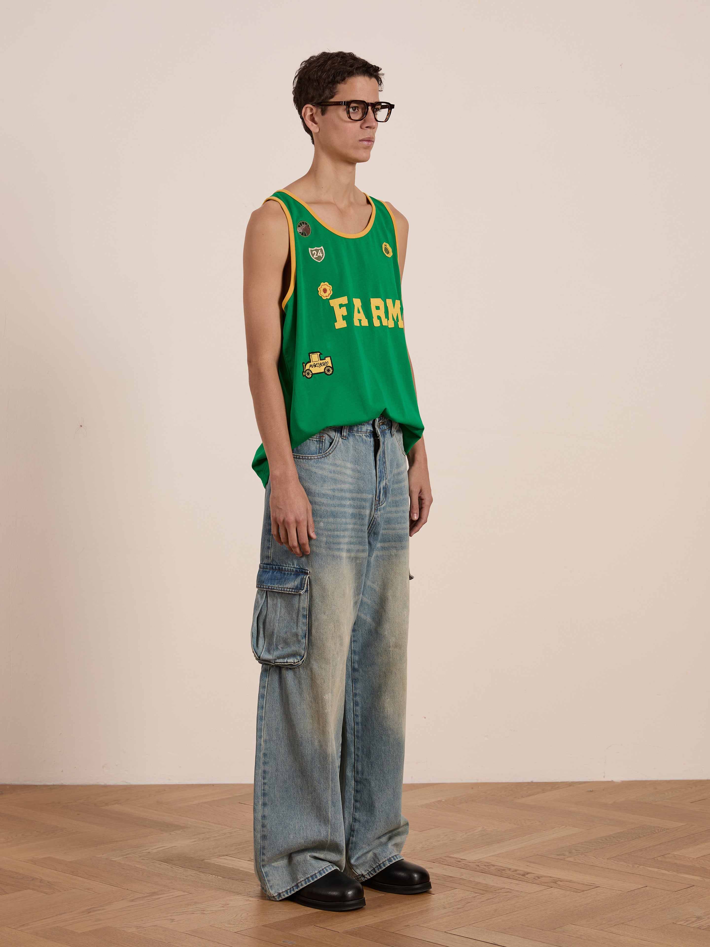 A person stands in profile, wearing FOUND's Farm Tank glasses and a green tank top with embroidered "FARM" patches. The vintage athletic vibe is completed with wide-leg jeans featuring cargo pockets. The wooden floor enhances the rustic charm of the look.