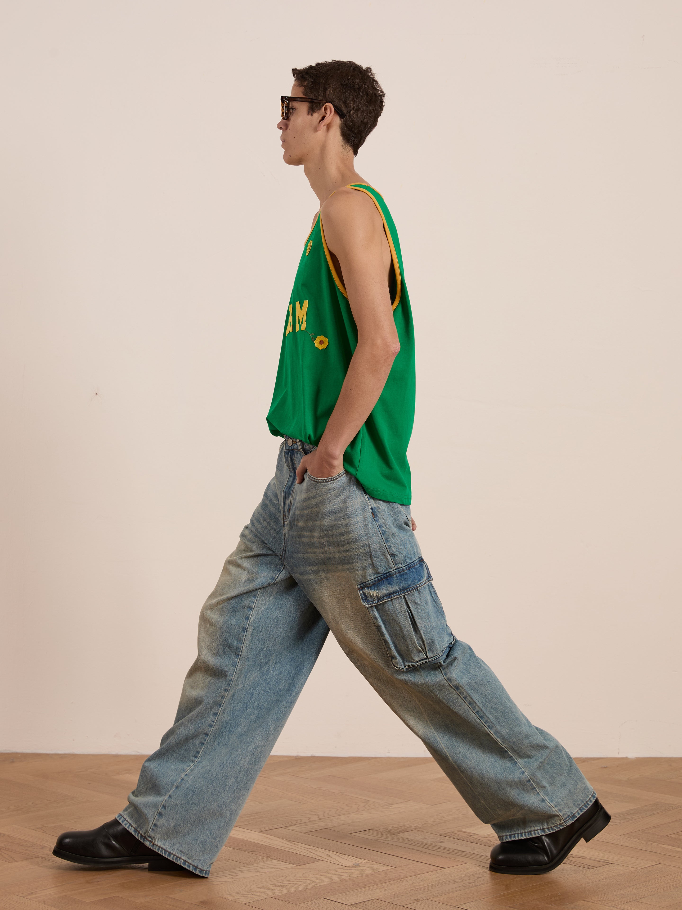 Wearing FOUND's green Farm Tank jersey made from breathable cotton and baggy jeans with embroidered patches, complemented by black shoes and sunglasses, a person strolls confidently on a wooden floor.