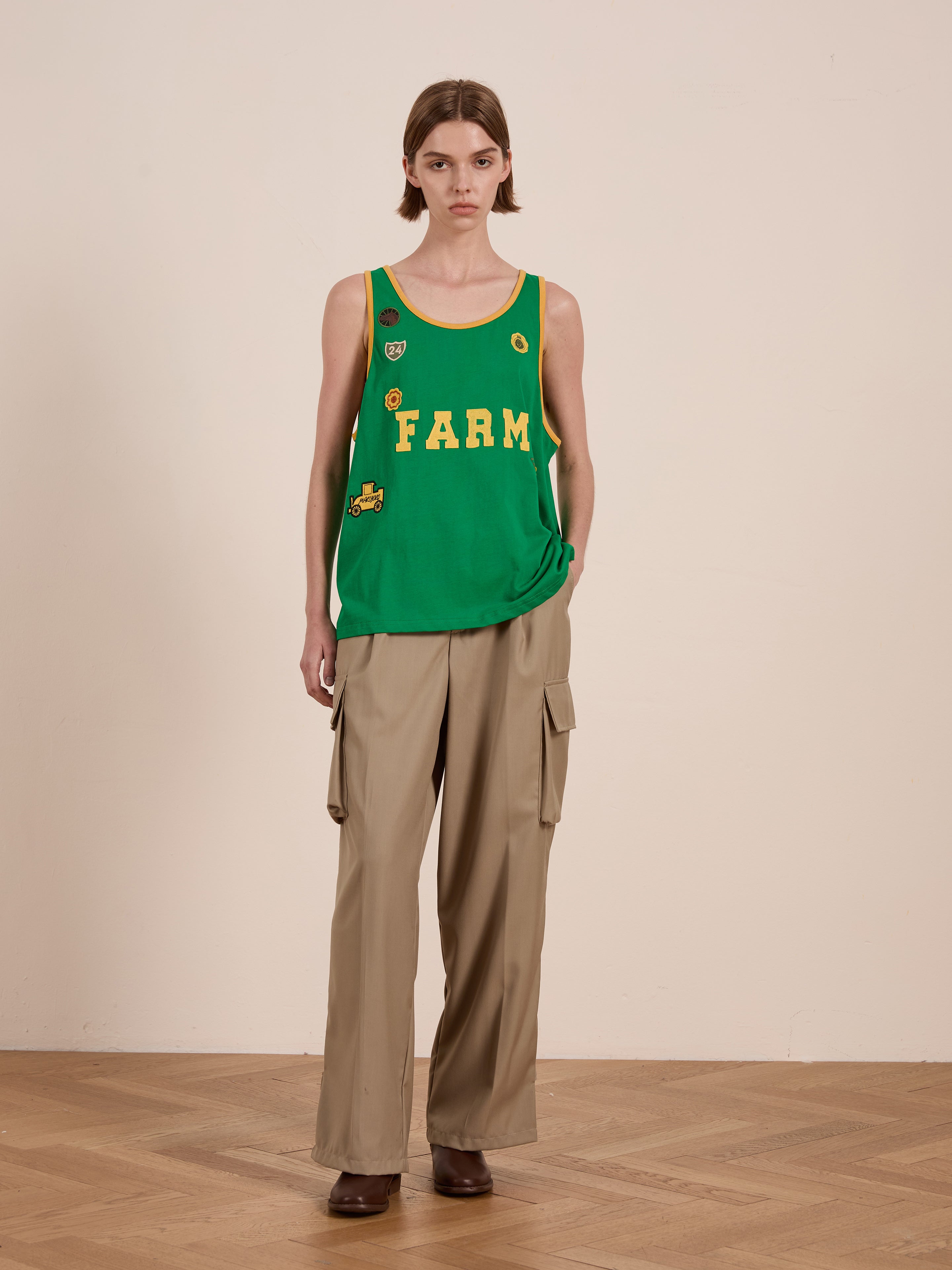 A person wears FOUND's Farm Tank, a green tank top with "FARM" text, paired with beige cargo pants reminiscent of vintage athletic wear. They stand on wooden flooring, their look finished with brown shoes against a plain background.