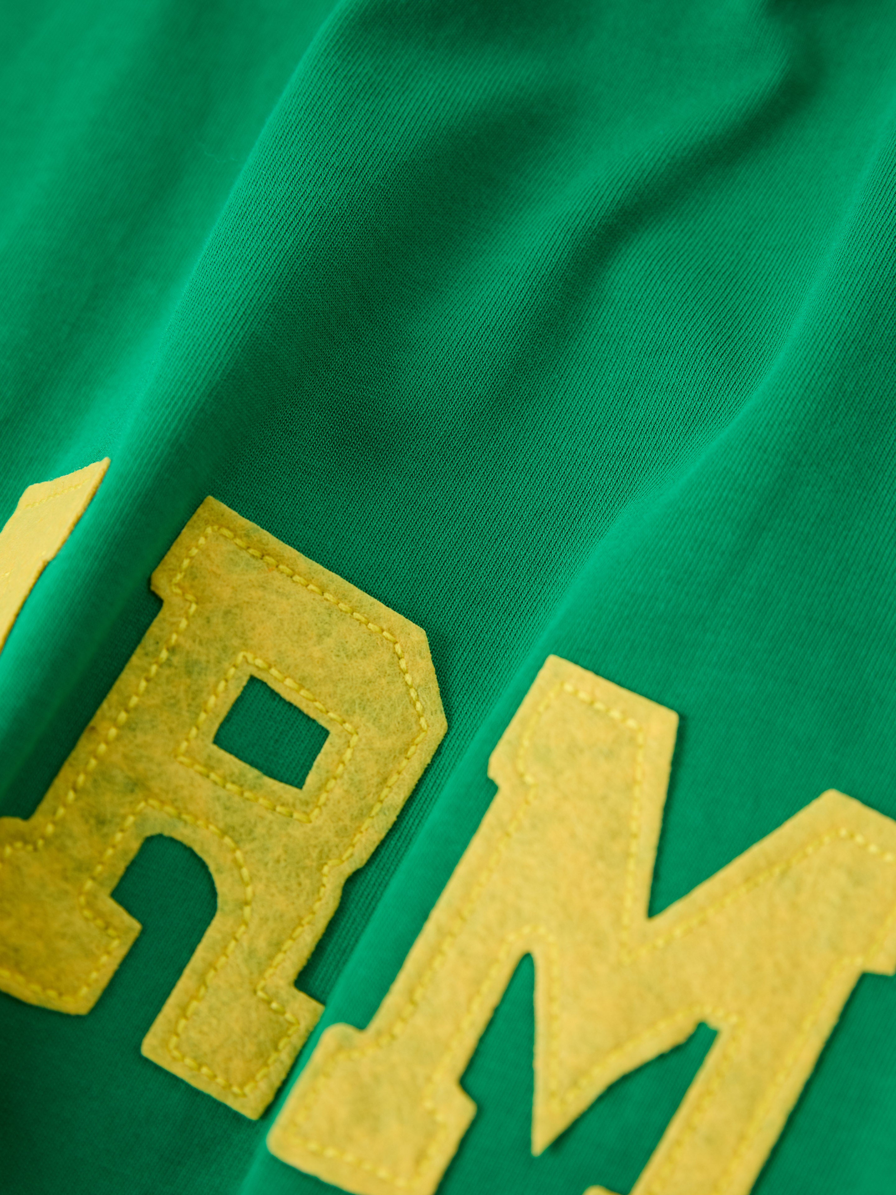 Close-up of a green Farm Tank by FOUND, featuring large yellow felt letters "R" and "M" reminiscent of vintage athletic wear.