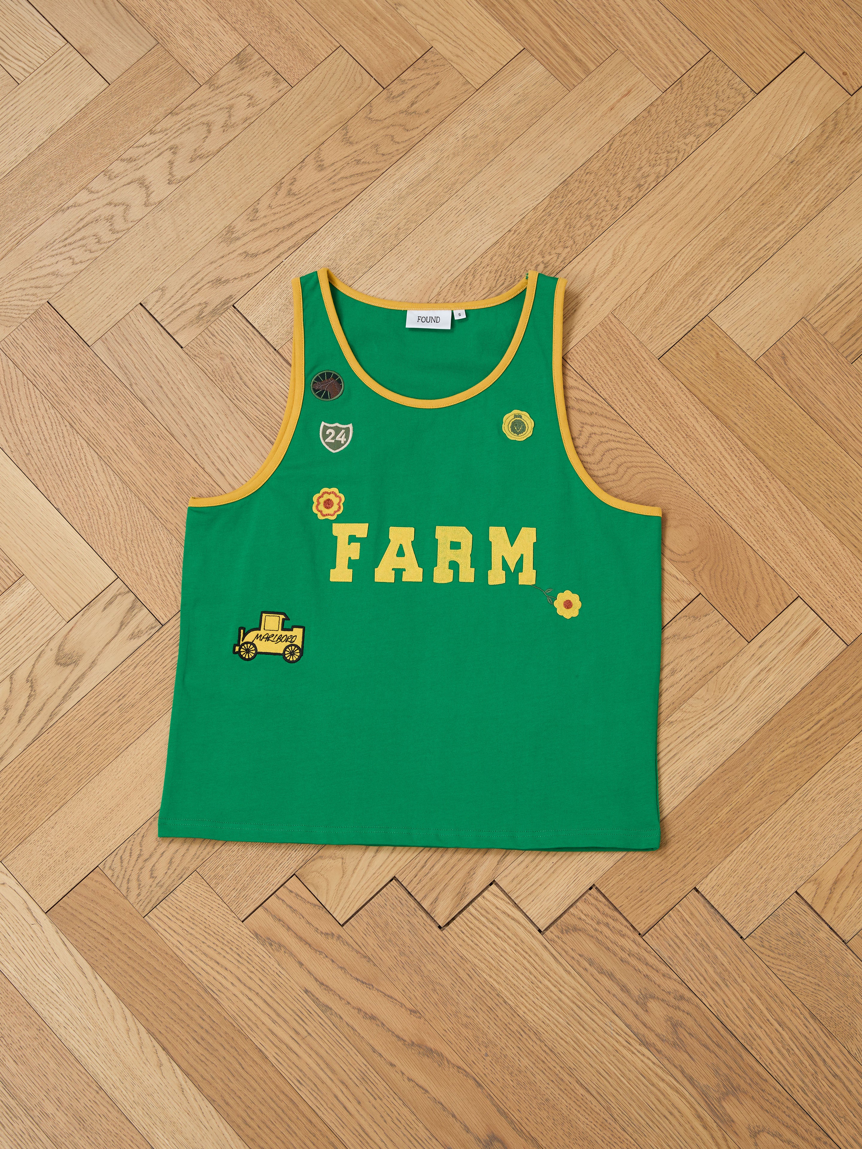 The Farm Tank by FOUND is a green tank top made from breathable cotton, featuring "FARM" in yellow, embroidered patches like a tractor, and exudes vintage athletic flair on a herringbone wooden floor.