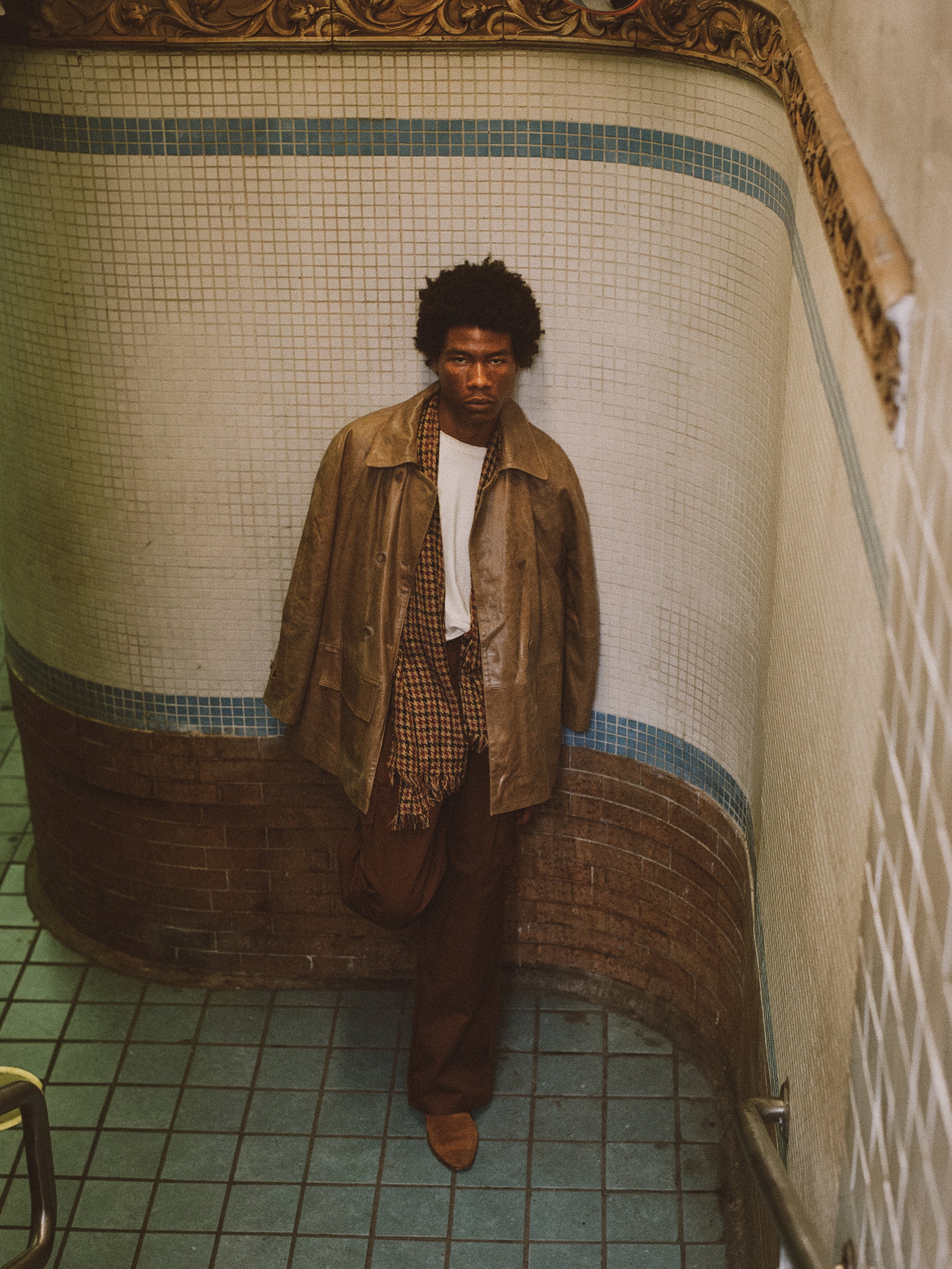 A person with curly hair leans against a tiled wall at the bottom of a staircase, wearing FOUND's ethically produced, vintage-inspired Fawn Double Breasted Leather Jacket, paired with a plaid shirt and brown pants.