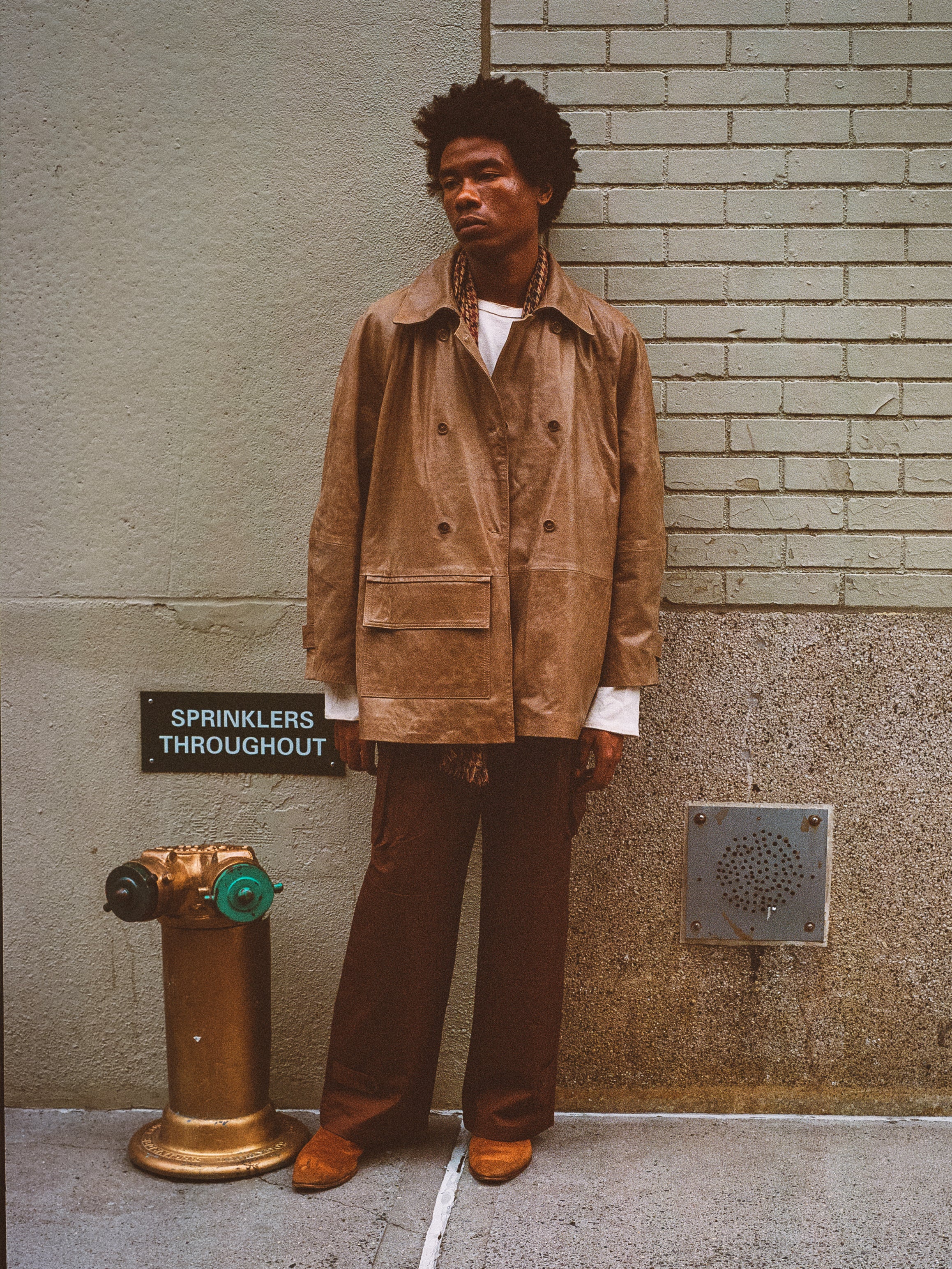 A person in FOUND's vintage-inspired Fawn Double-Breasted Leather Jacket leans against a brick wall next to a fire hydrant, creating an effortlessly stylish look with their brown pants.