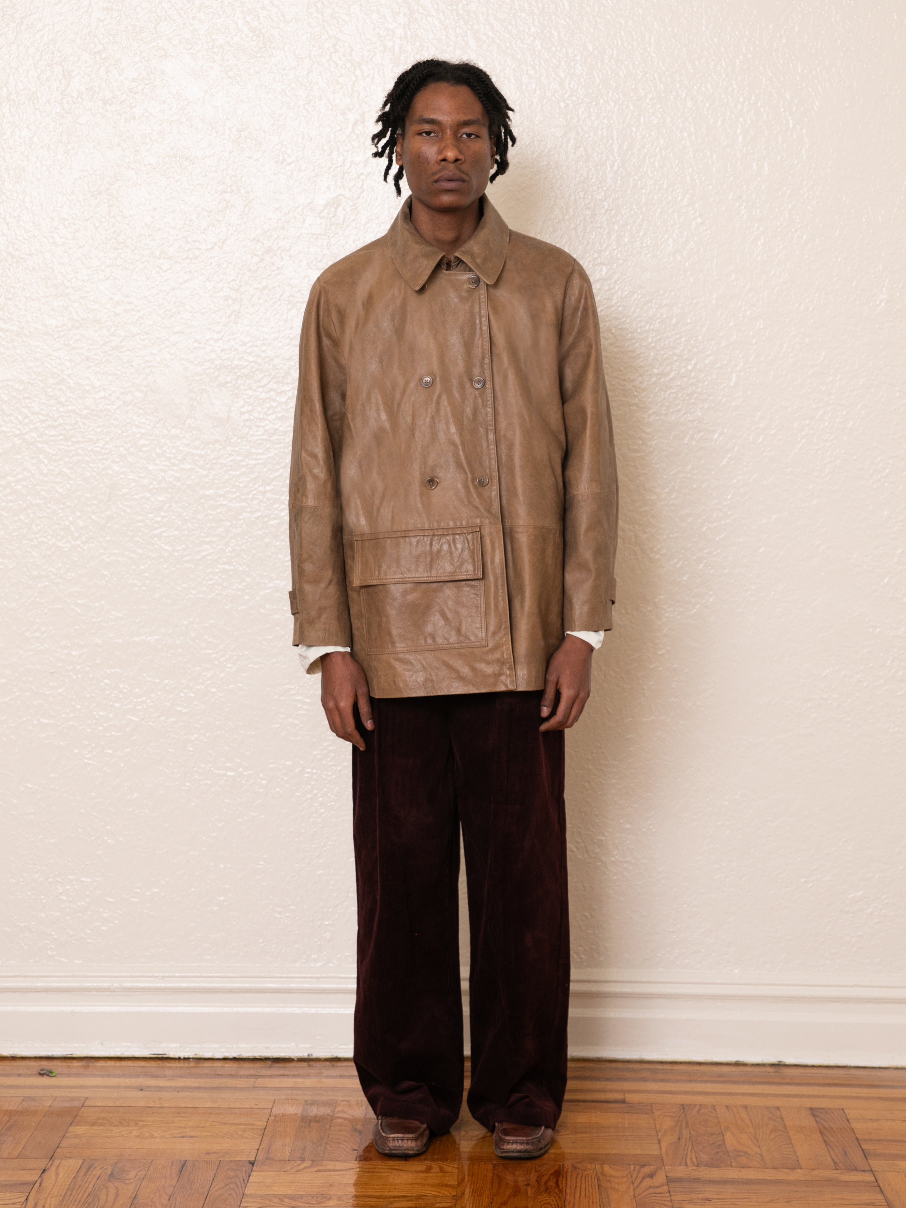 A person wearing a FOUND Fawn Double Breasted Leather Jacket and dark pants stands against a light-colored wall on a wooden floor.