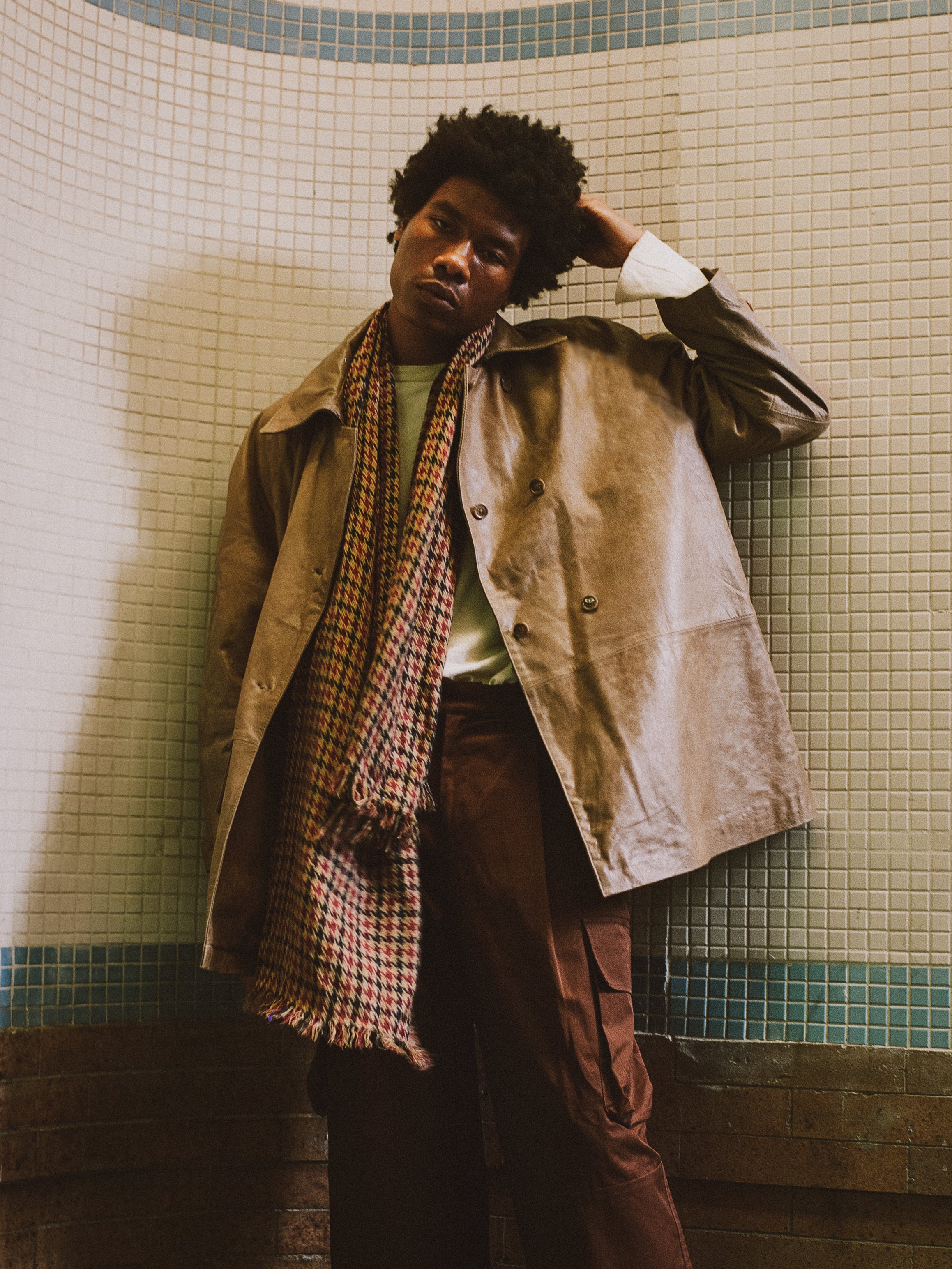 A person in a FOUND Fawn Double Breasted Leather Jacket, a checkered scarf, and brown pants stands against a tiled wall, gently touching their head.