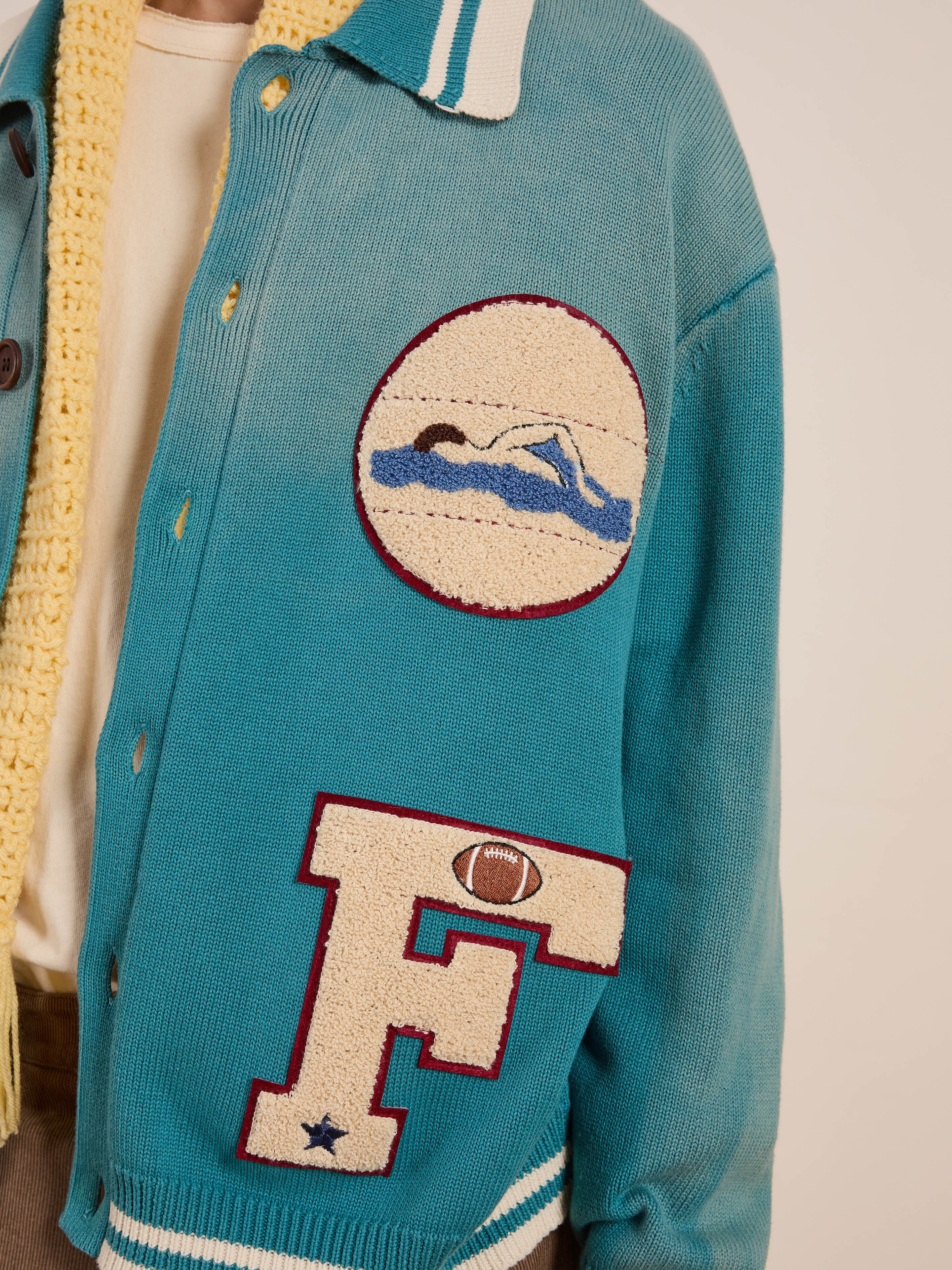 A person sports the FOUND Fin Varsity Patch Collared Cardigan, a vintage-style blue varsity jacket featuring a swimming patch, a letter "F" with a football emblem, and a yellow collar, perfecting the collegiate look.