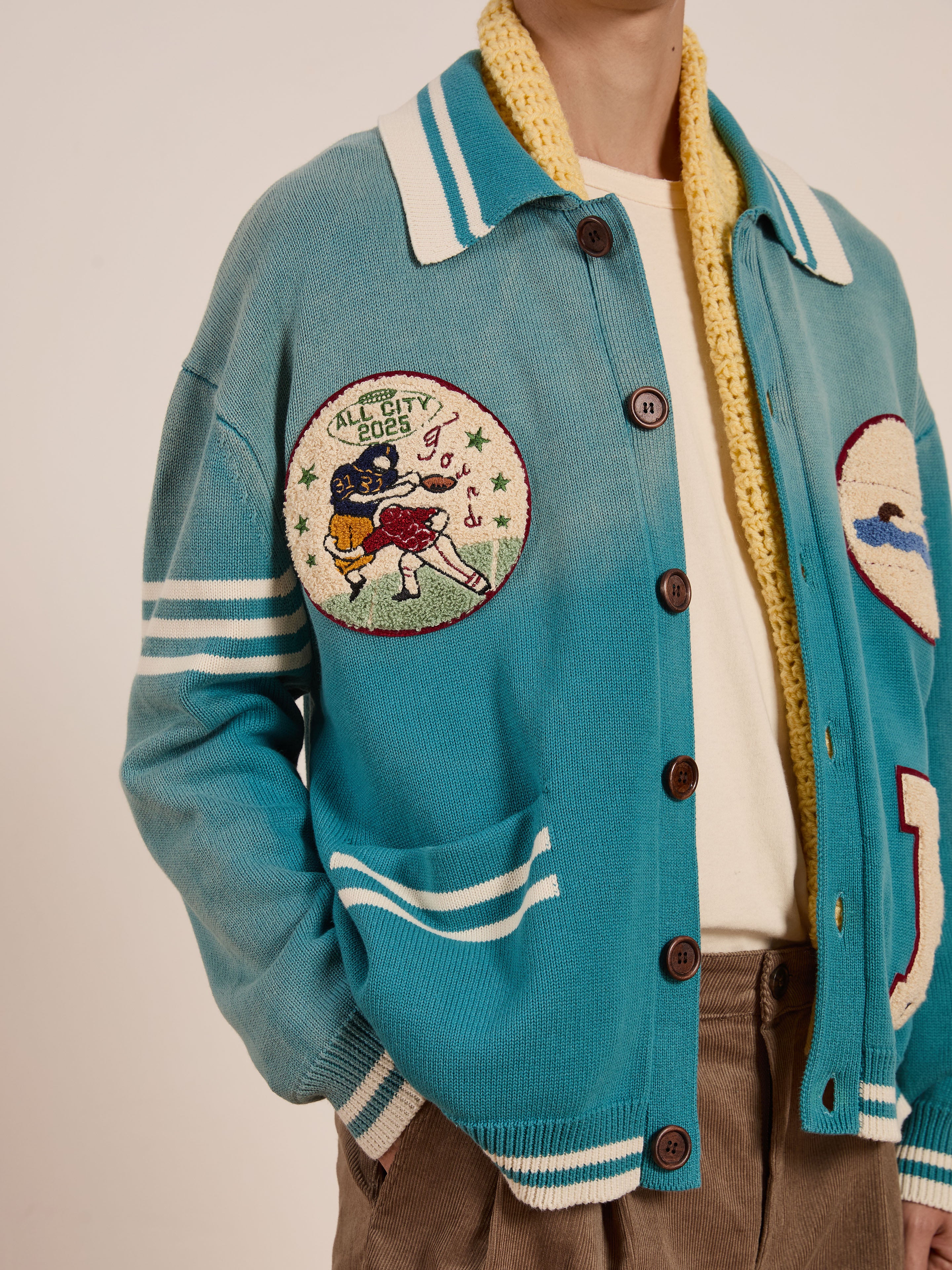 A person wears FOUND's Fin Varsity Patch Collared Cardigan in blue, featuring an embroidered sports patch with "All City 2023," exuding a vintage collegiate style.